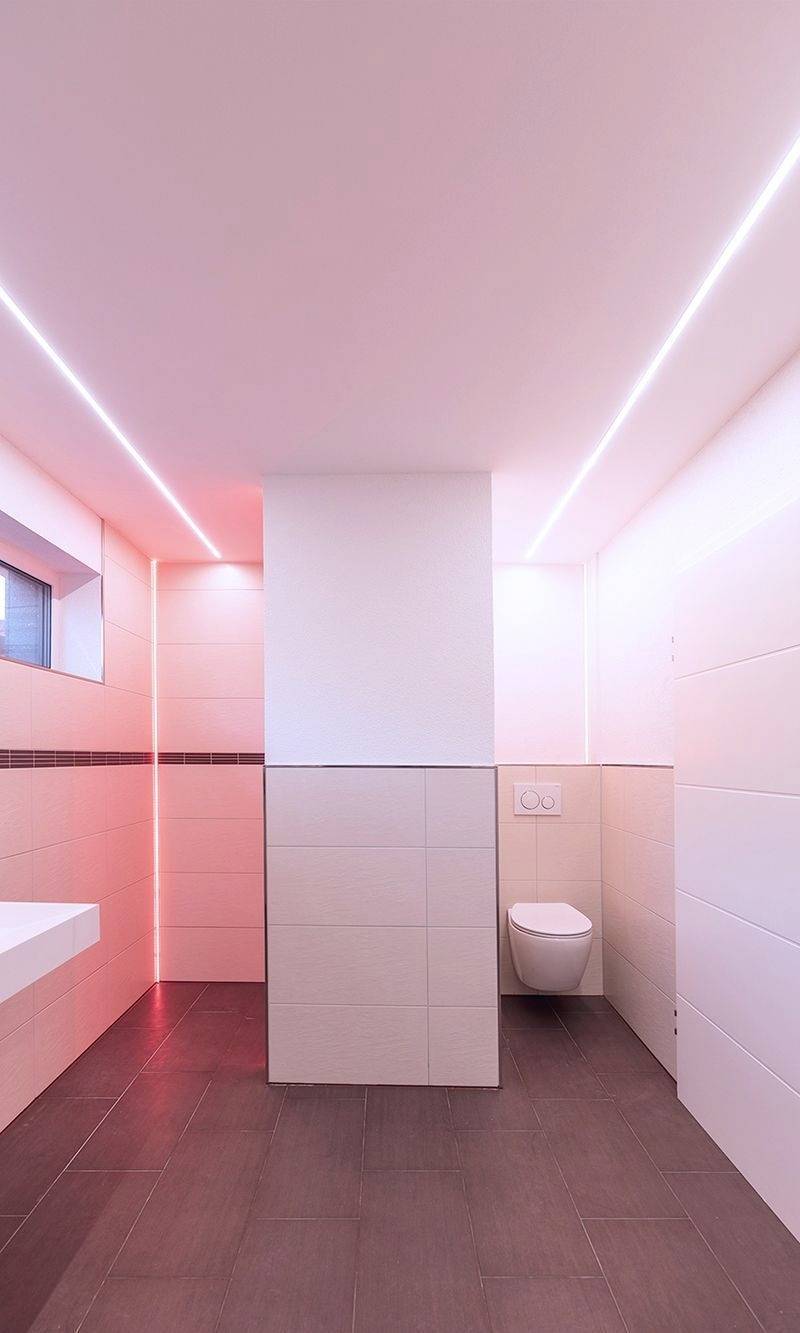 Modernt designat badrum med indirekt belysning, rosa och vita kakel. Väggmonterad toalett och handfat finns.