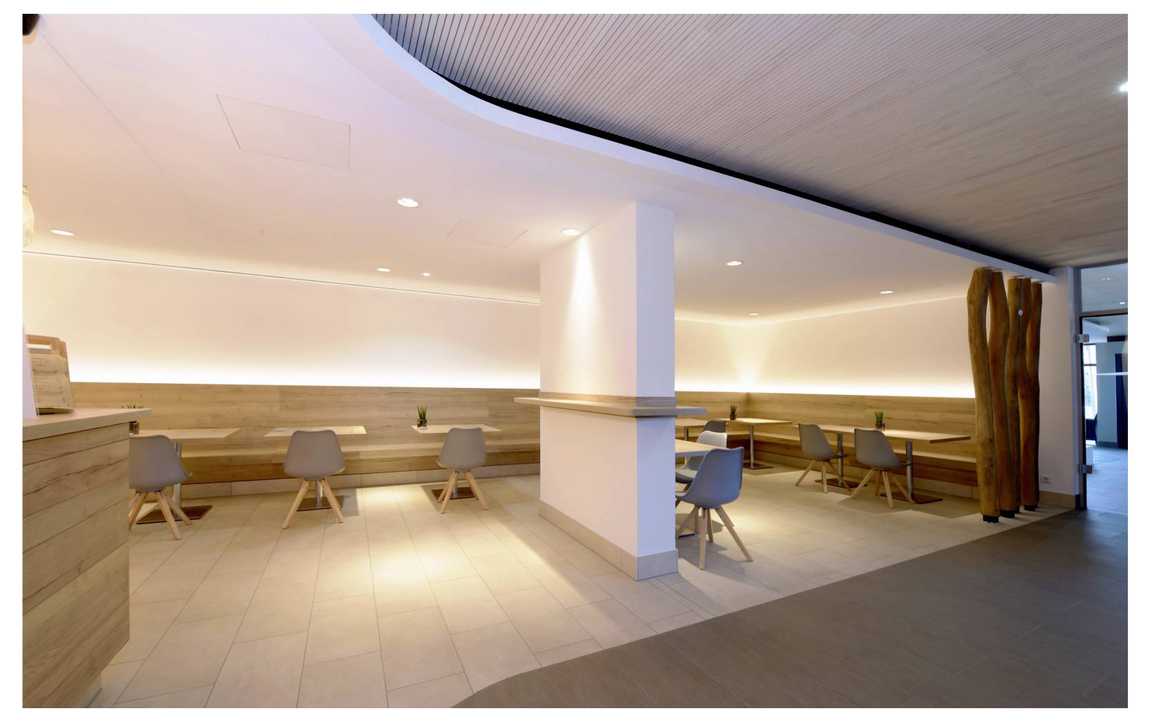Modern minimalistisk café-interiör med träbänkar, småbord och stolar, en central pelare, varm infälld belysning och ett öppet sittområde.