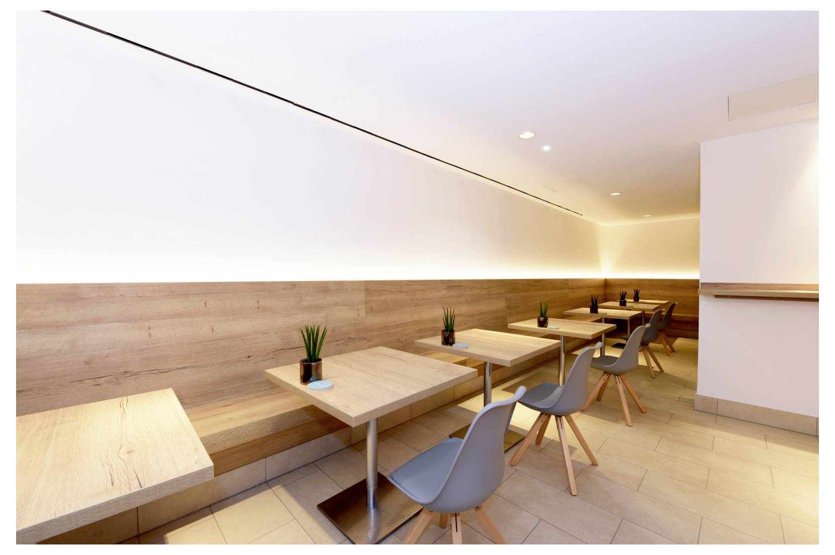 Modern minimalistisk café-interiör med tomma träbord, grå stolar och varmt väggbelysning längs en lång bänk