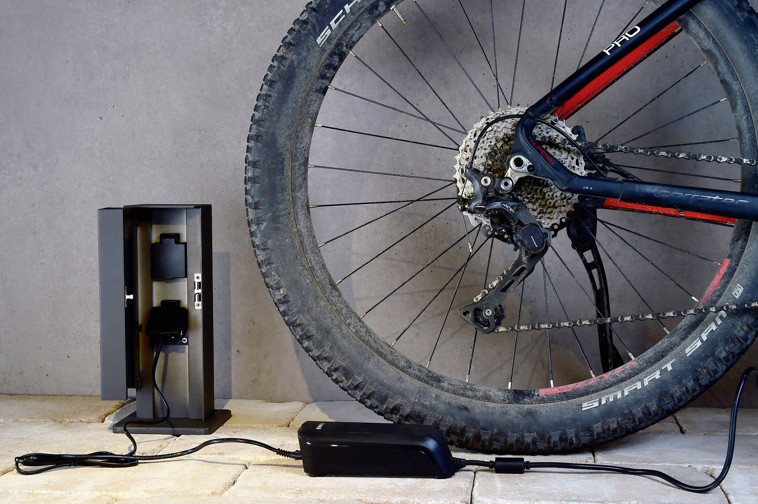 En cykel står vid en vägg, bakhjulet visas. Bredvid finns en laddningsstation med ett isatt batteri.