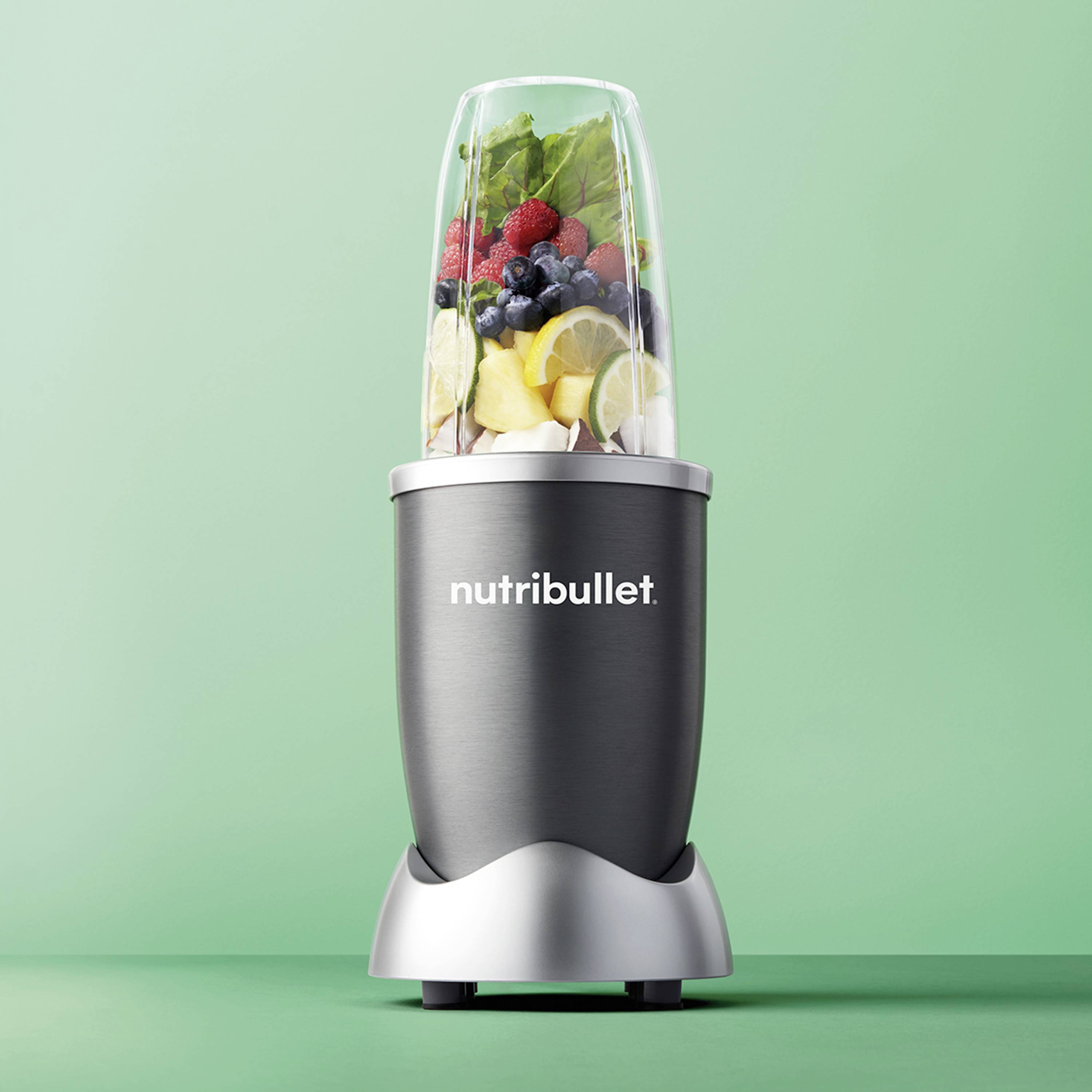 En grå mixer fylld med frukt och grönsaker, inklusive bär och citronskivor, framför en grön bakgrund. Texten 'nutribullet' på apparaten.