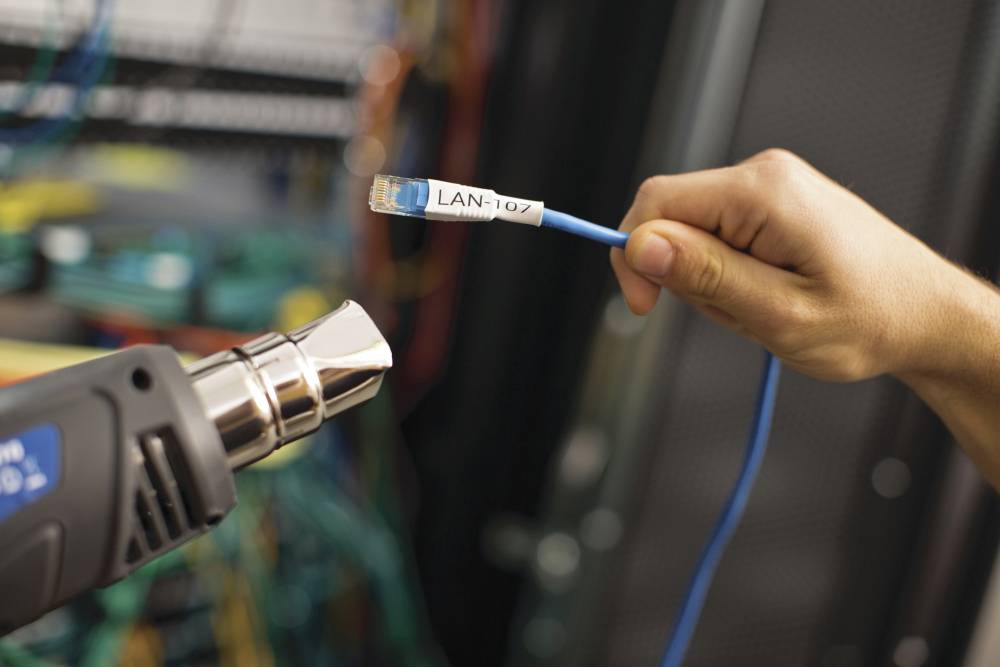 En person håller en LAN-kabel framför en etikettskrivare i ett serverrum. Kabeln är märkt med 'LAN-107'.