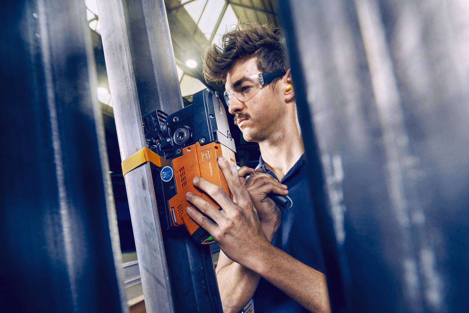 En man med skyddsglasögon kontrollerar strukturen på en metallbalk med ett mätinstrument i en industriell miljö.