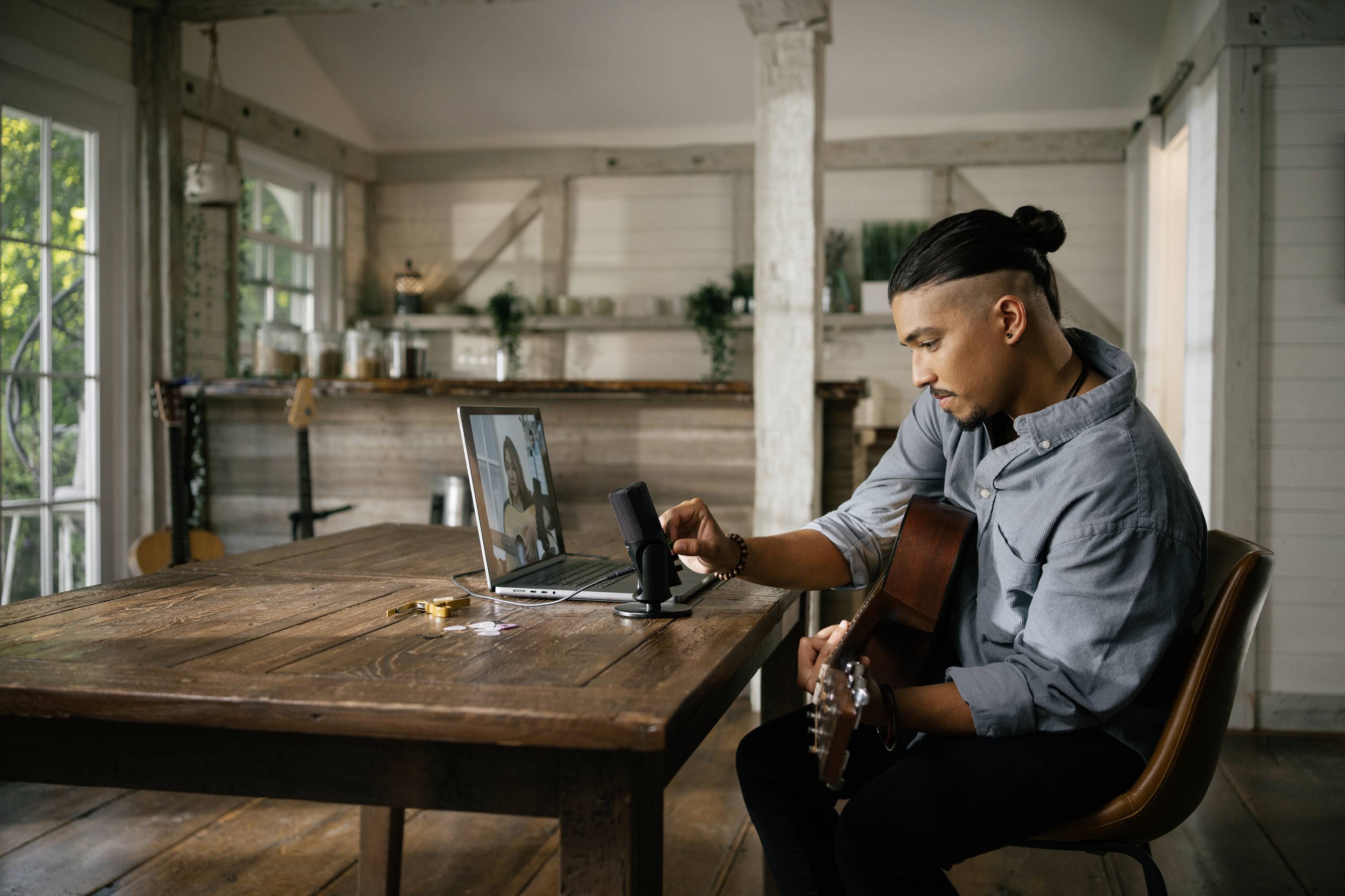 En person sitter vid ett bord med en gitarr och tittar på en smartphone som är placerad framför en bärbar dator. Rummet är hemtrevligt möblerat.