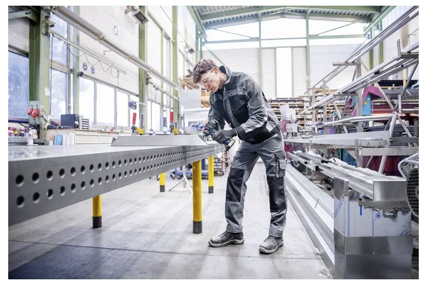 En arbetare i skyddsutrustning använder ett handverktyg för att inspektera eller montera en stor metallbalk inuti en industriell fabrik.