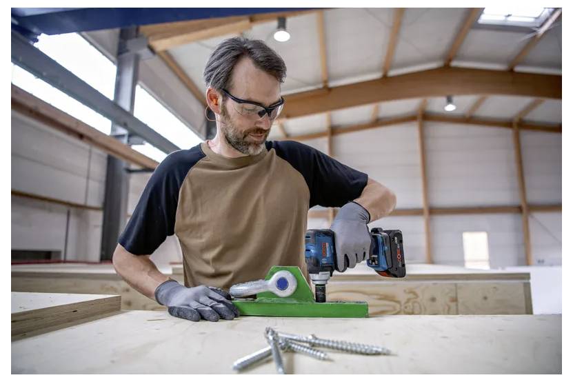 En man som bär skyddsglasögon och handskar använder en eldriven borrmaskin på ett grönt metallstycke i en träverkstad med synliga bjälkar och skruvar.