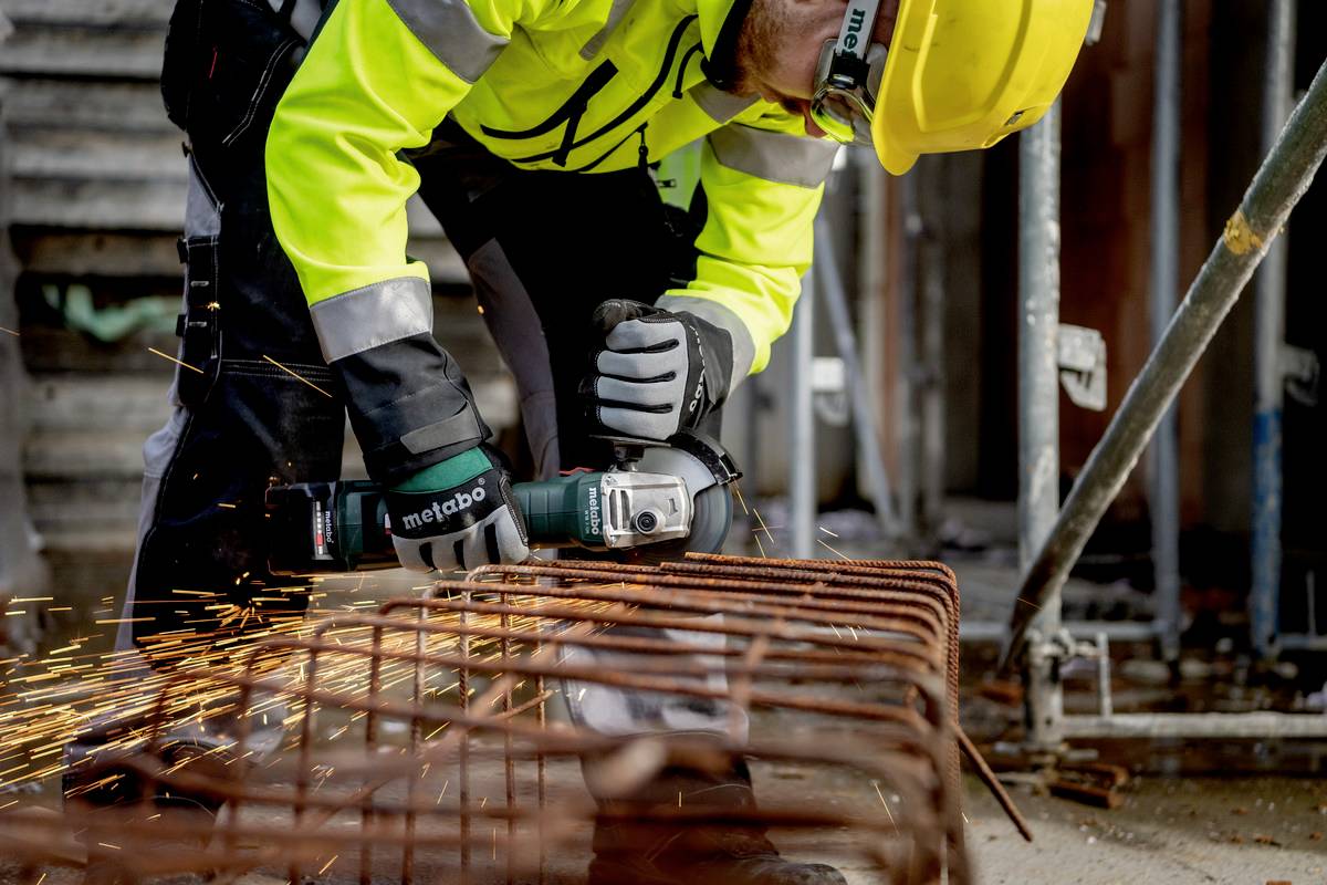 En byggnadsarbetare i skyddsutrustning klipper stålstänger med en vinkelslip. Gnistor flyger från metallen. I bakgrunden syns en byggarbetsplats.