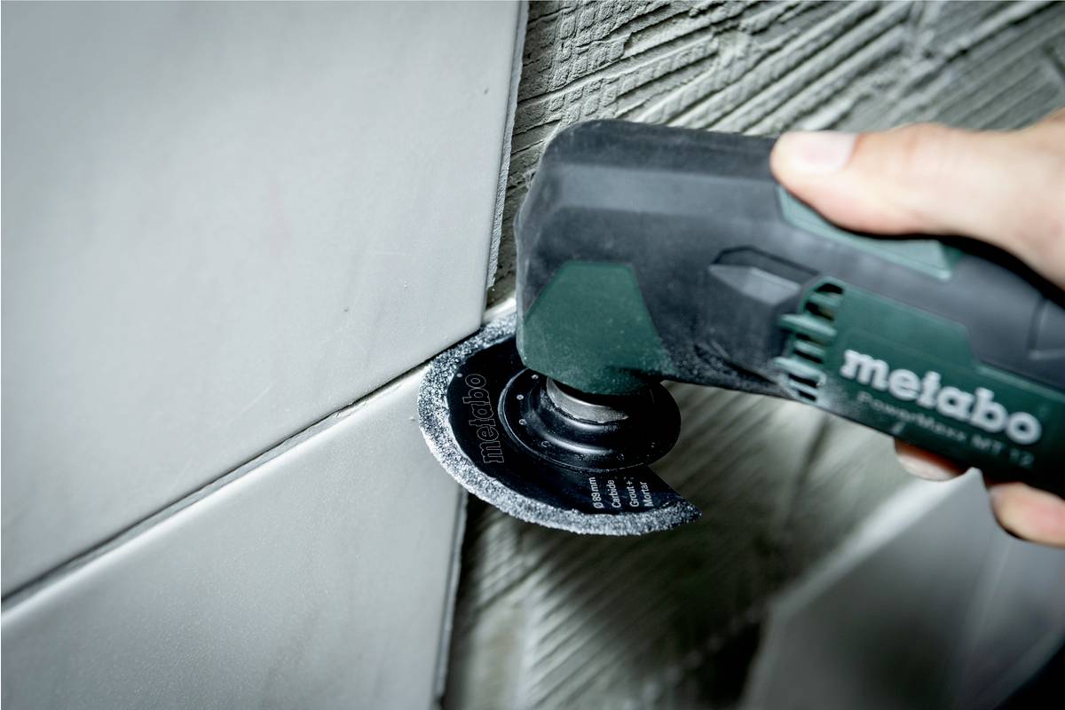 Hand håller elektriskt verktyg med liten såg, klipper fogar mellan väggplattor. Ytan av plattor och fogar synlig.
