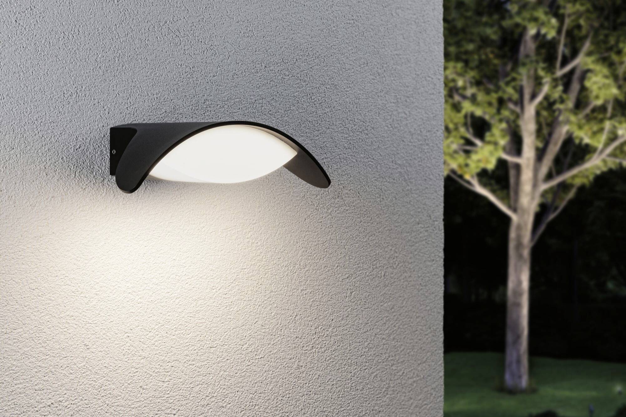 Vägglampa på en grå vägg utomhus, lyser upp omgivningen. Bakgrunden visar ett träd under natten.