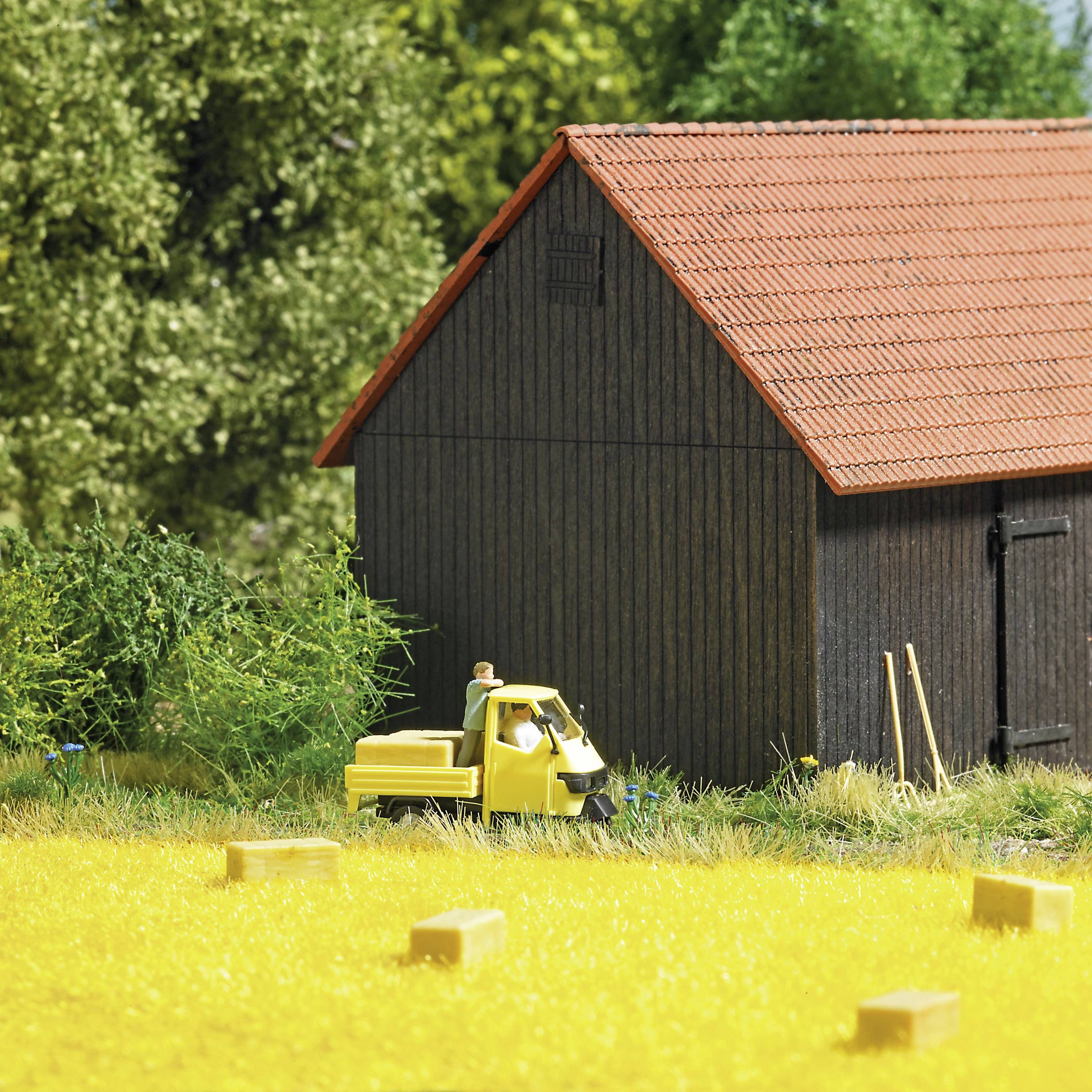 Ett gult leksaksbilar står framför en lada i ett modellandskap. I förgrunden ligger ett fält med höbalar, omgivet av träd.