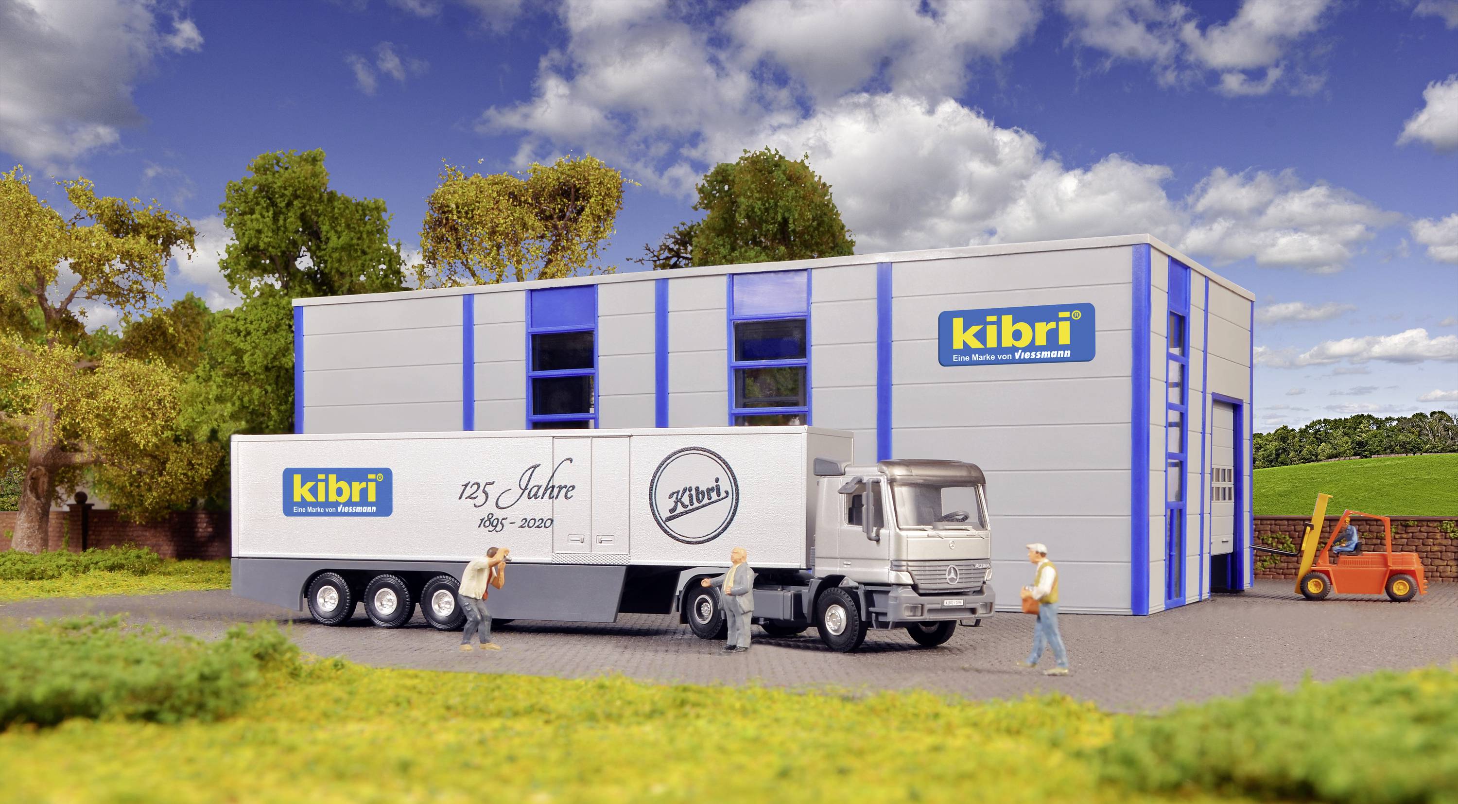 En modell-lastbil med inskriptionen '125 år' står framför ett byggnadscomplex med 'kibri' logotyp. Tre figurer och en gaffeltruck syns.