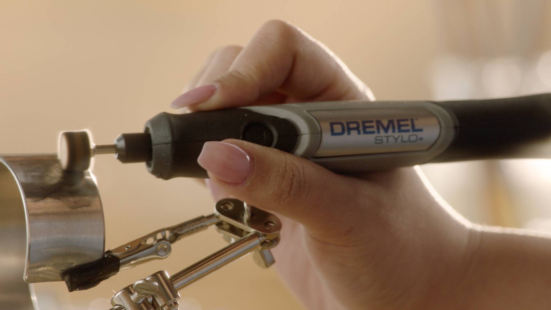 En hand håller ett Dremel-verktyg och bearbetar ett metallföremål som är fastsatt i ett skruvstäd.