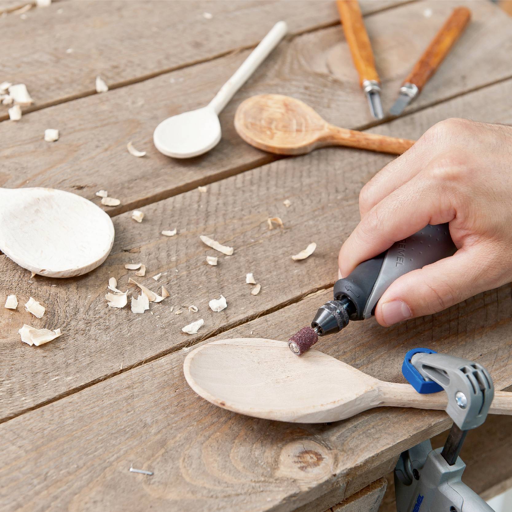 En hand bearbetar en träsked med ett Dremel-verktyg på ett träbord. Träspån och ytterligare skedar syns i bilden.