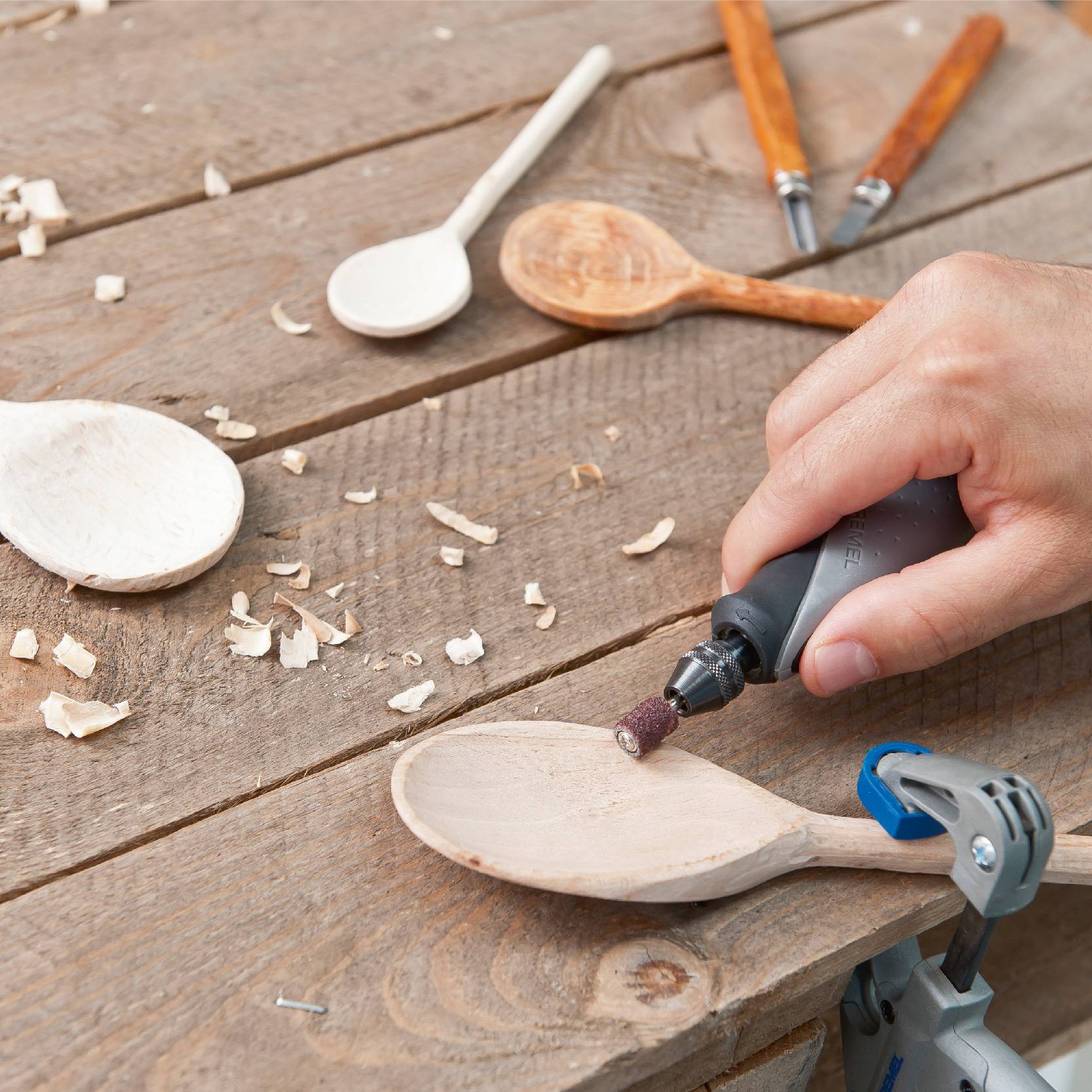 En hand slipar en träsked med ett Dremel-verktyg, medan flera träspån och andra skedar ligger på bordet.