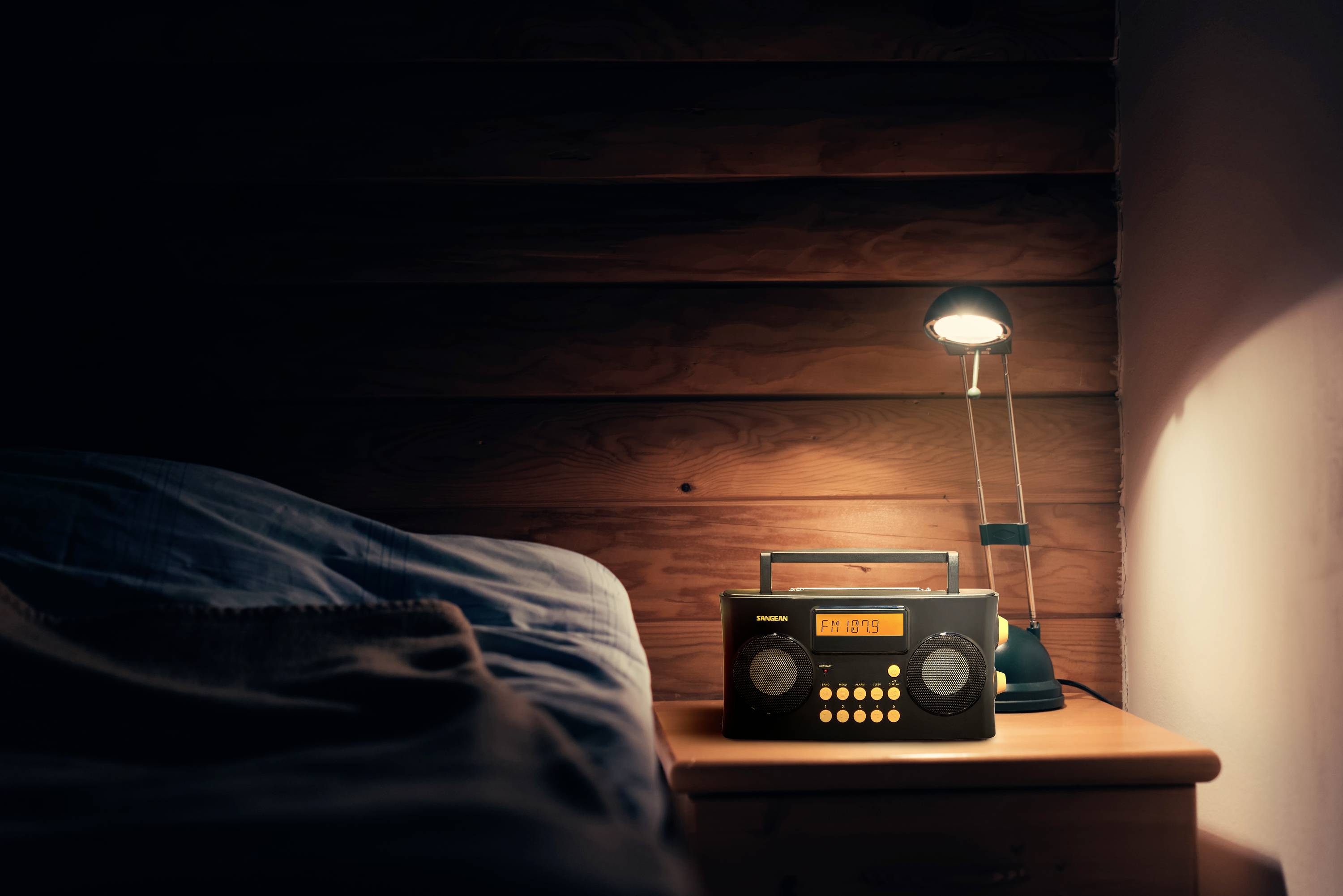 En nattsängsradio och en lampa på ett träbord bredvid en säng. Lampan lyser mjukt upp det mörka träet och radion.