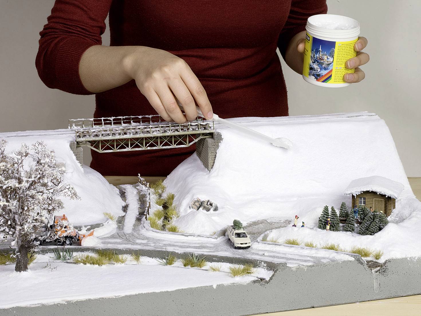 En person formar en miniatyrmiljö på vintern, strör vitt pulver som modelsnö. En bro och en bil är synliga.