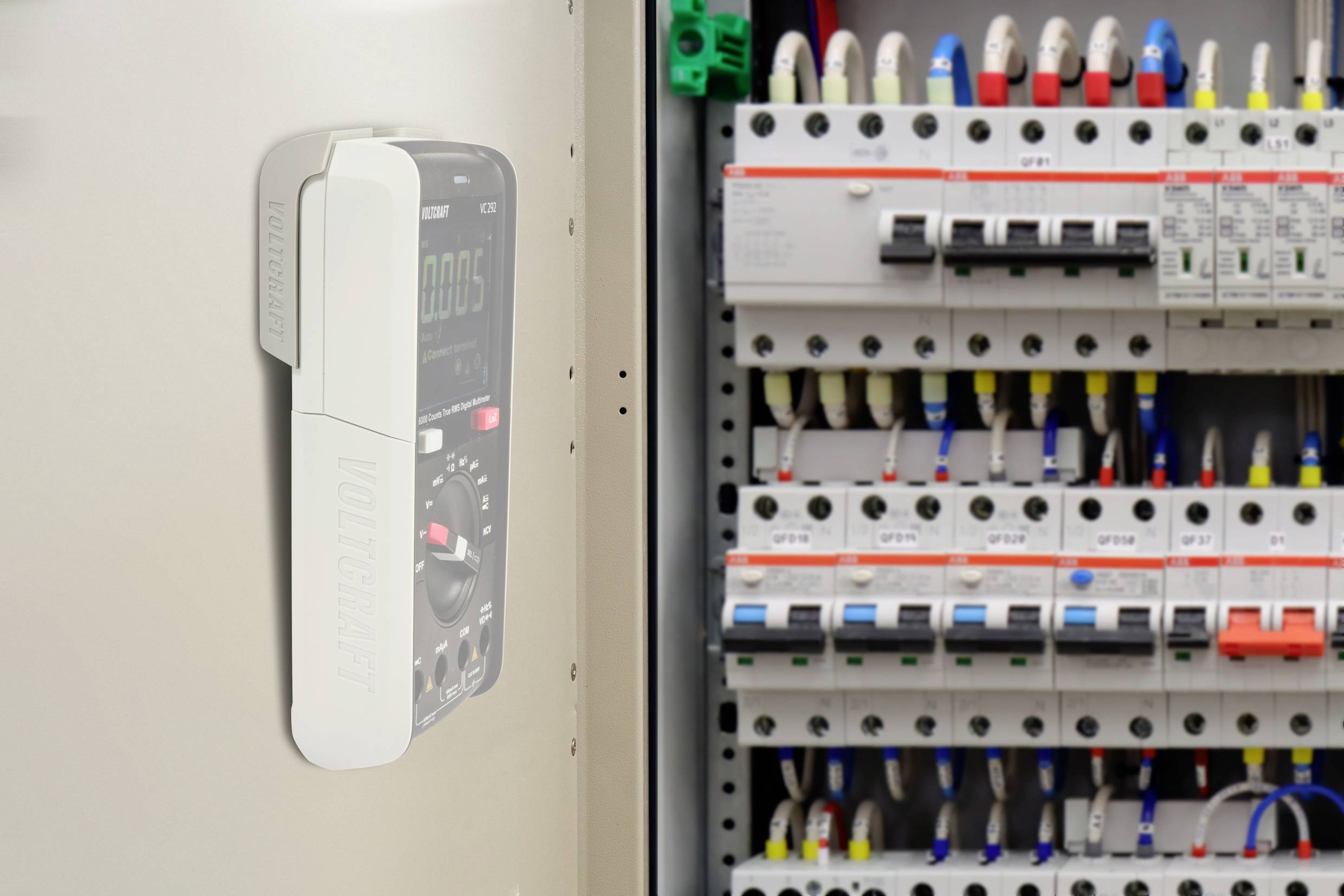En multimeter mäter spänning i en elcentral full av säkringar och strömbrytare. Paneler med olika färgmarkeringar synliga.