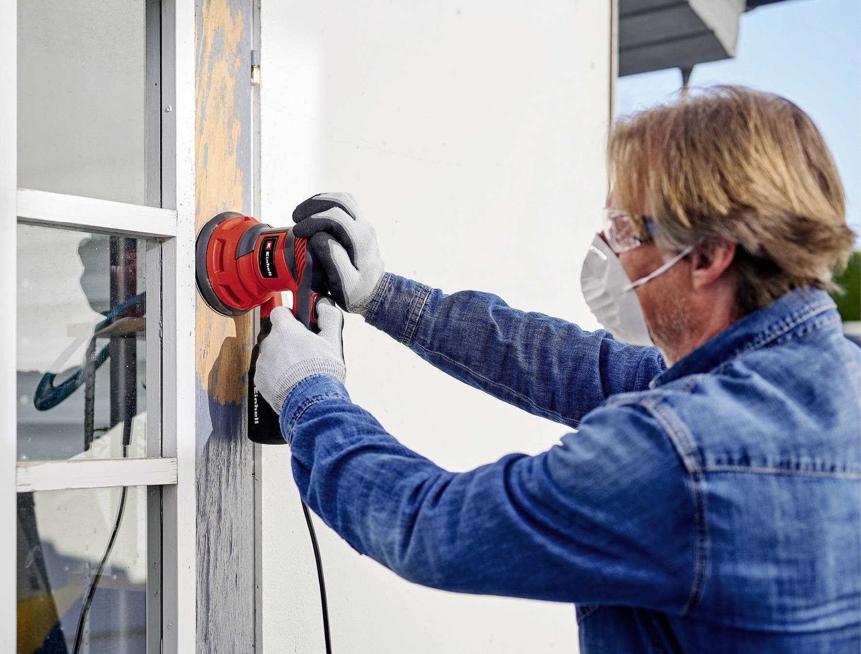 En man slipar träkarmen till ett fönster med en elektrisk slipmaskin. Han bär skyddsglasögon och mask.