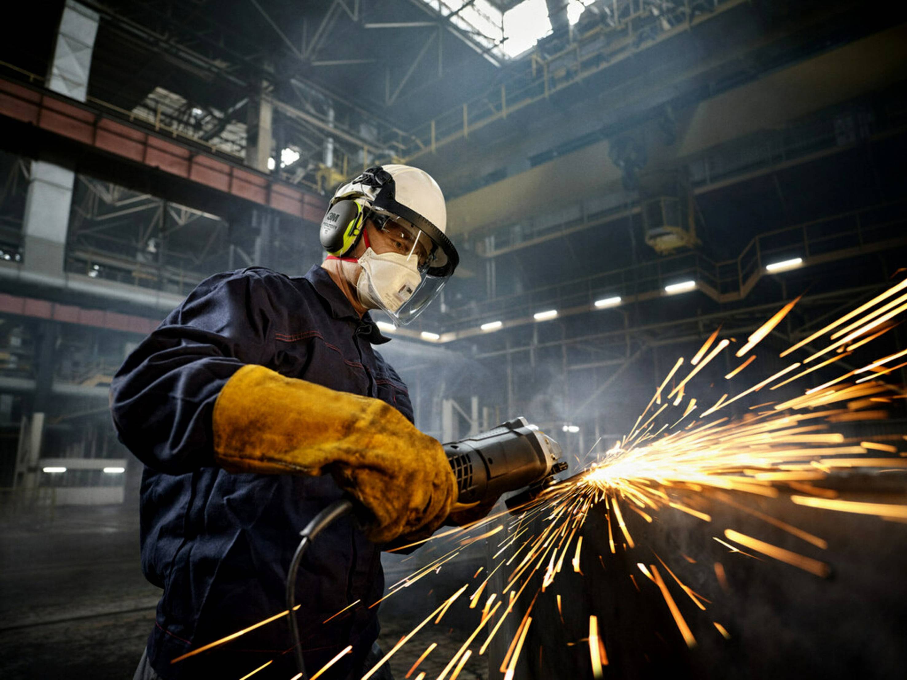 En arbetare i skyddsutrustning slipar metall i en fabrikshall. Gnistor flyger från slipmaskinens skiva.