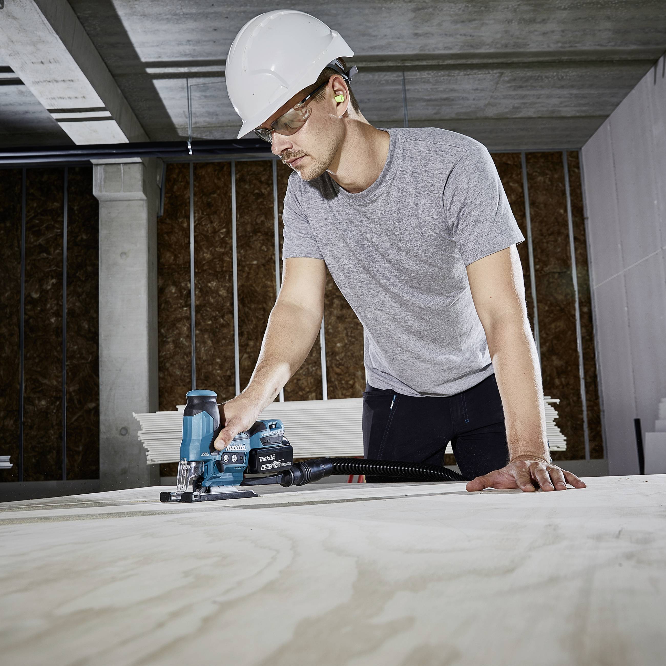 En man med skyddshjälm och hörselskydd använder ett elverktyg för att bearbeta trä i en industriell lokal.