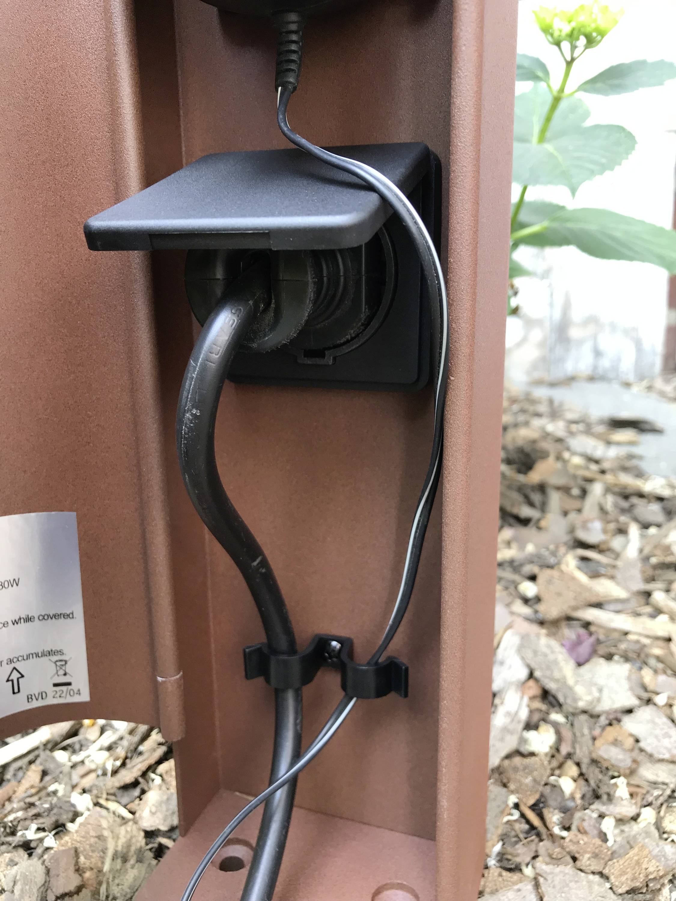 En utomhusuttag med öppet lock visar två isatta kablar. I bakgrunden syns växter och träflis.