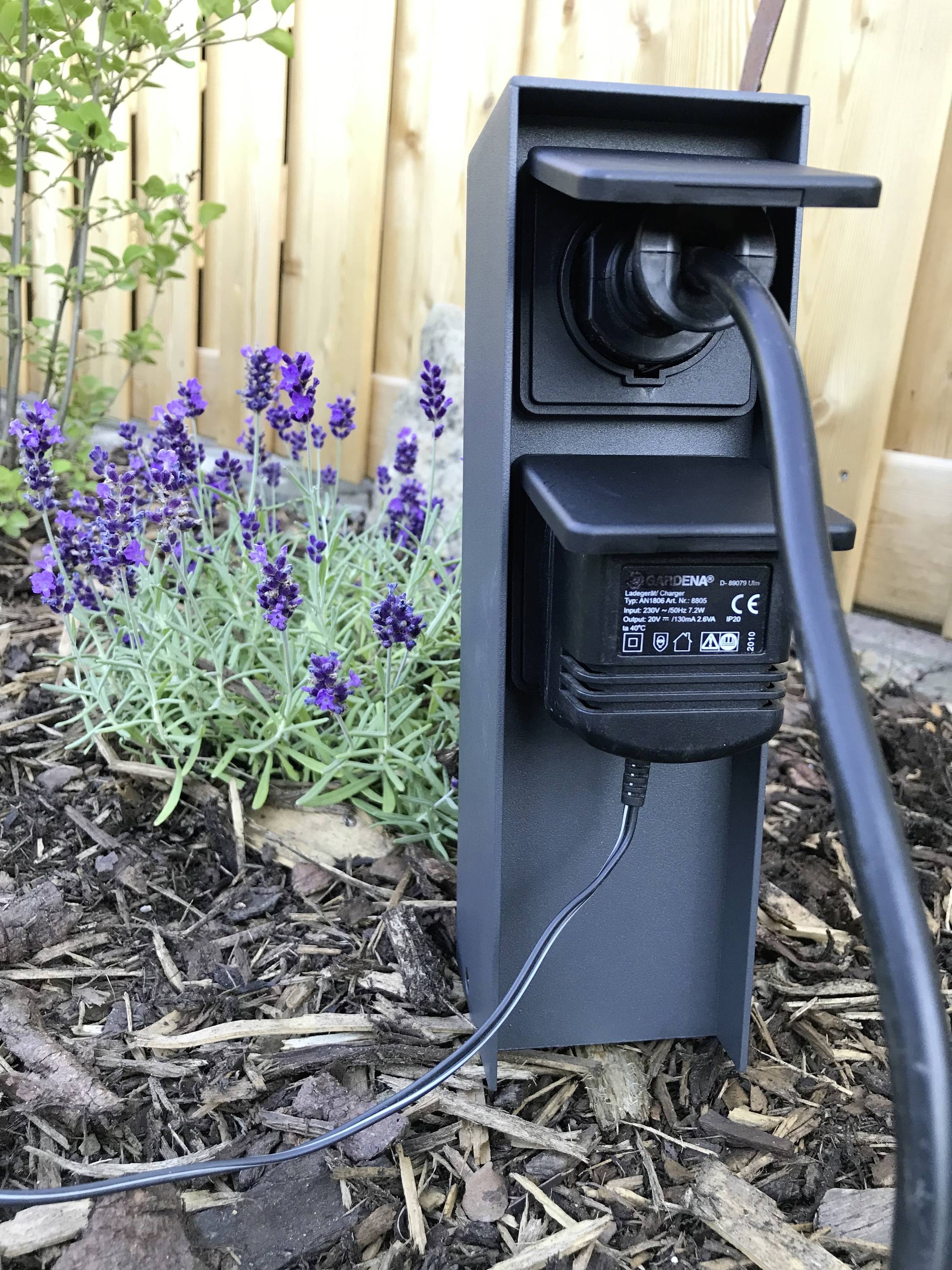 Ett eluttag utomhus med en ansluten kabel och blommor i bakgrunden.