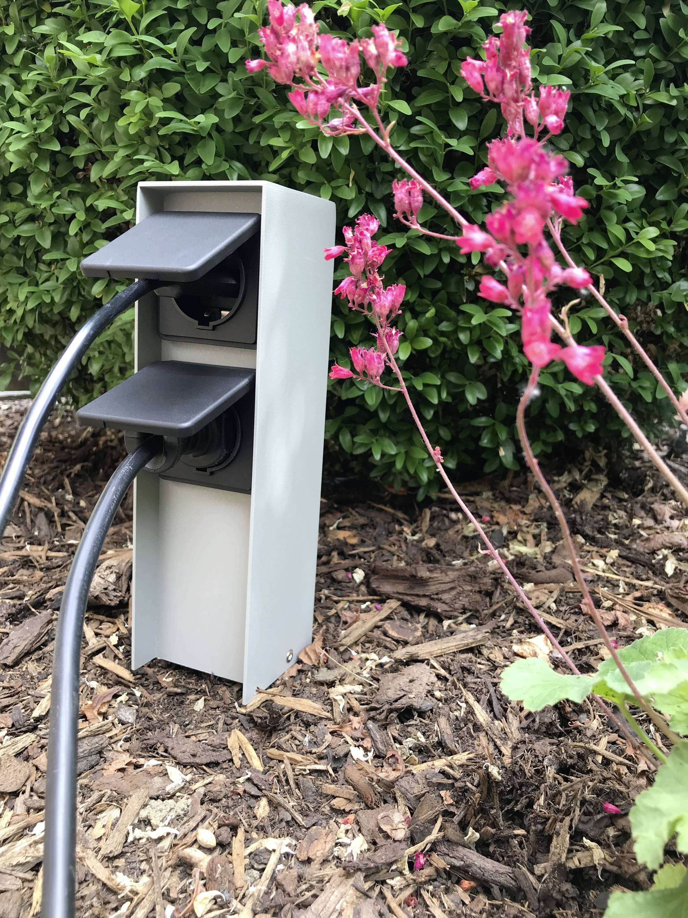 En utomhusuttag med två anslutna kablar finns i trädgården, omgiven av blommor och gröna löv.