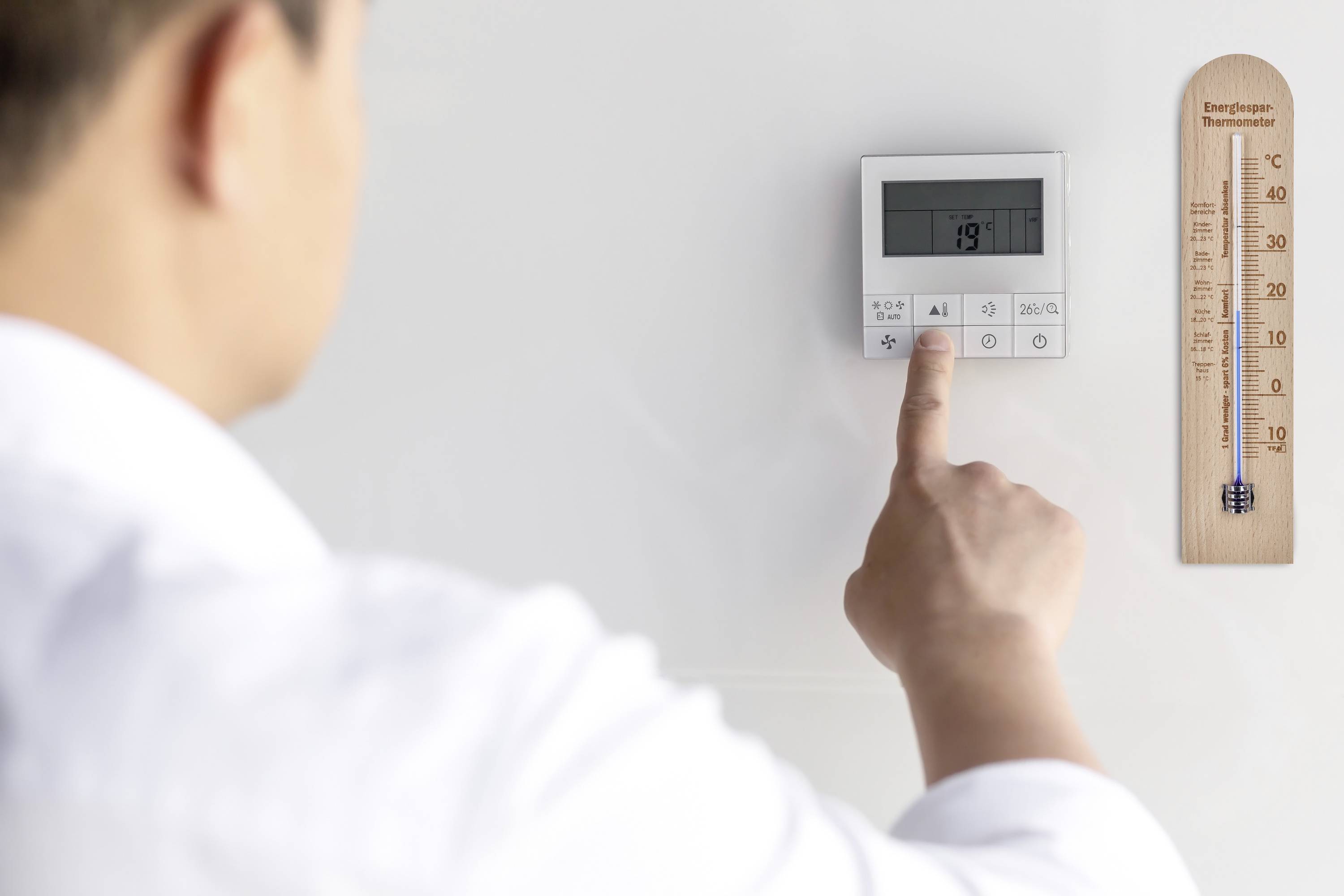 En person ställer in en digital termostat på 19 grader. Bredvid till höger visar ett träattrapp-termometer temperaturen i Celsius.