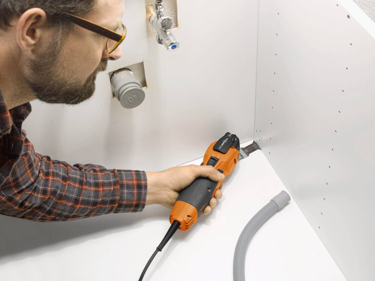 En man använder ett elektriskt multiverktyg för att bearbeta ett hörn i ett vitt träskåp. Slangar och rör är synliga.