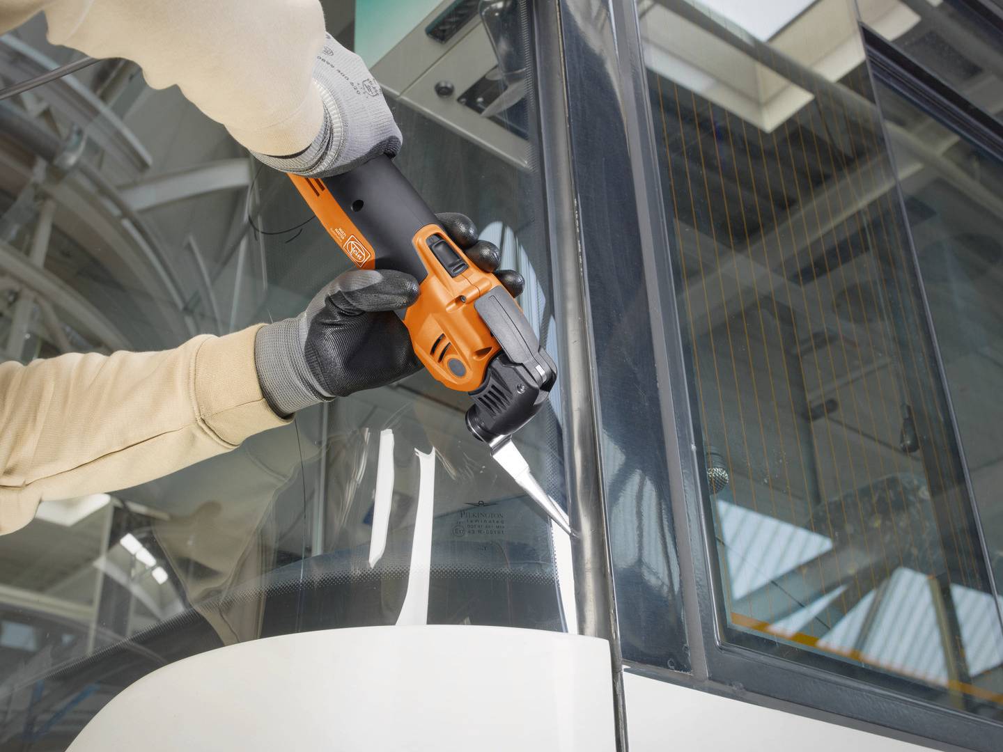 En person arbetar med ett orangefärgat elverktyg vid ett fordonsfönster. Verktyget klipper eller bearbetar glas.