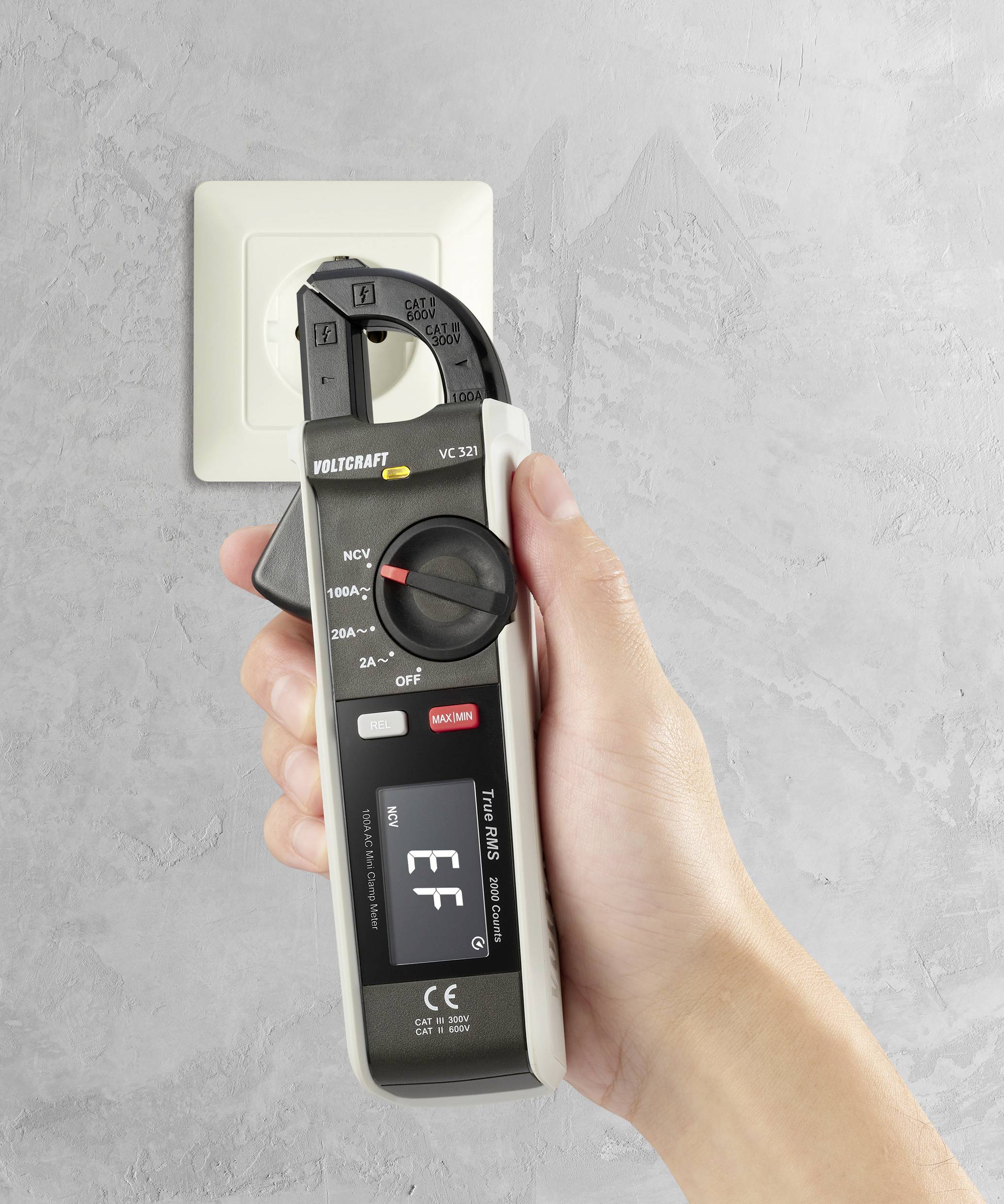 En hand håller ett spänningsmätinstrument framför ett eluttag för att kontrollera elektriska egenskaper.