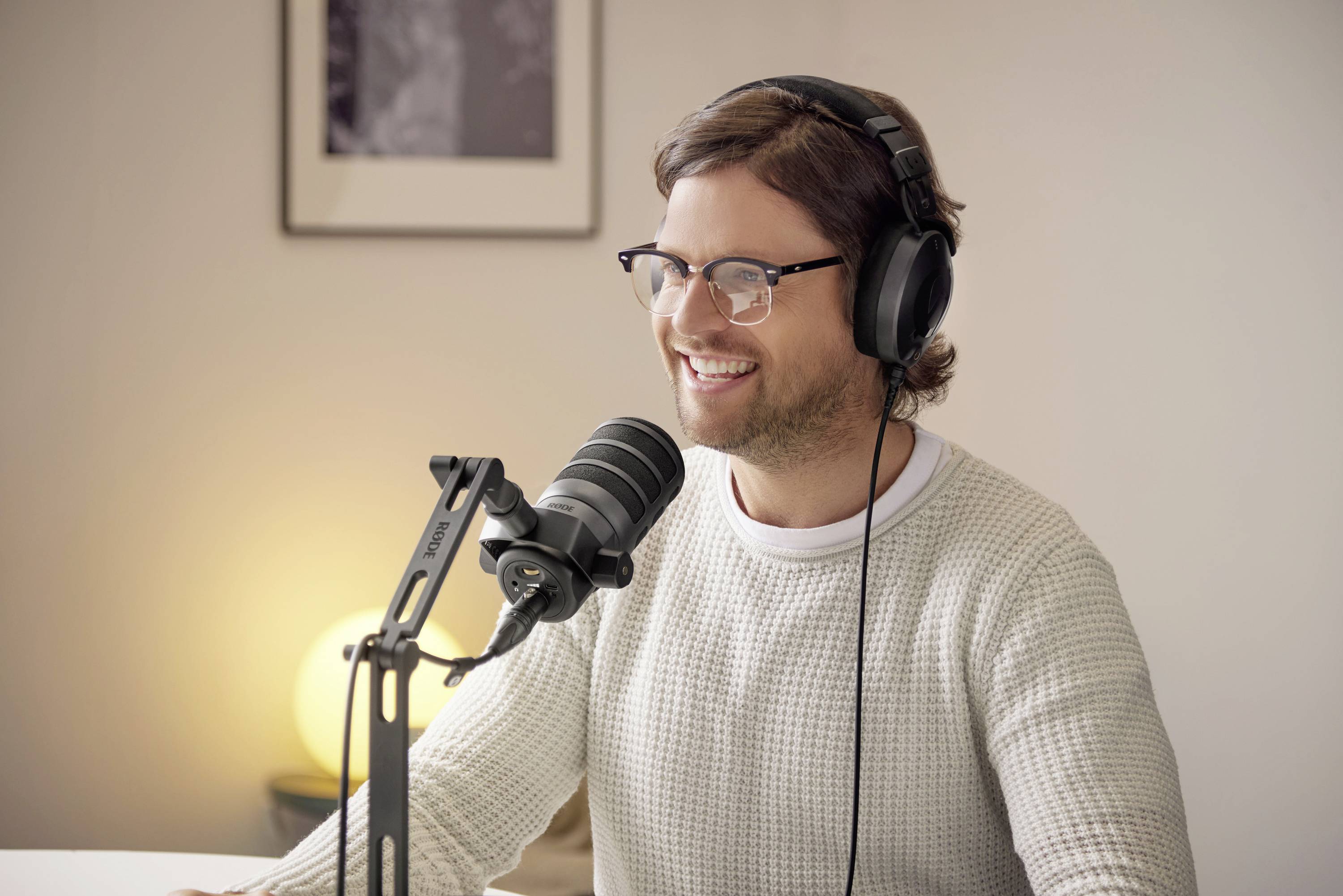 Person med hörlurar och mikrofon, sitter leende vid ett bord och deltar i en ljudinspelning eller podcast i ett mysigt rum.