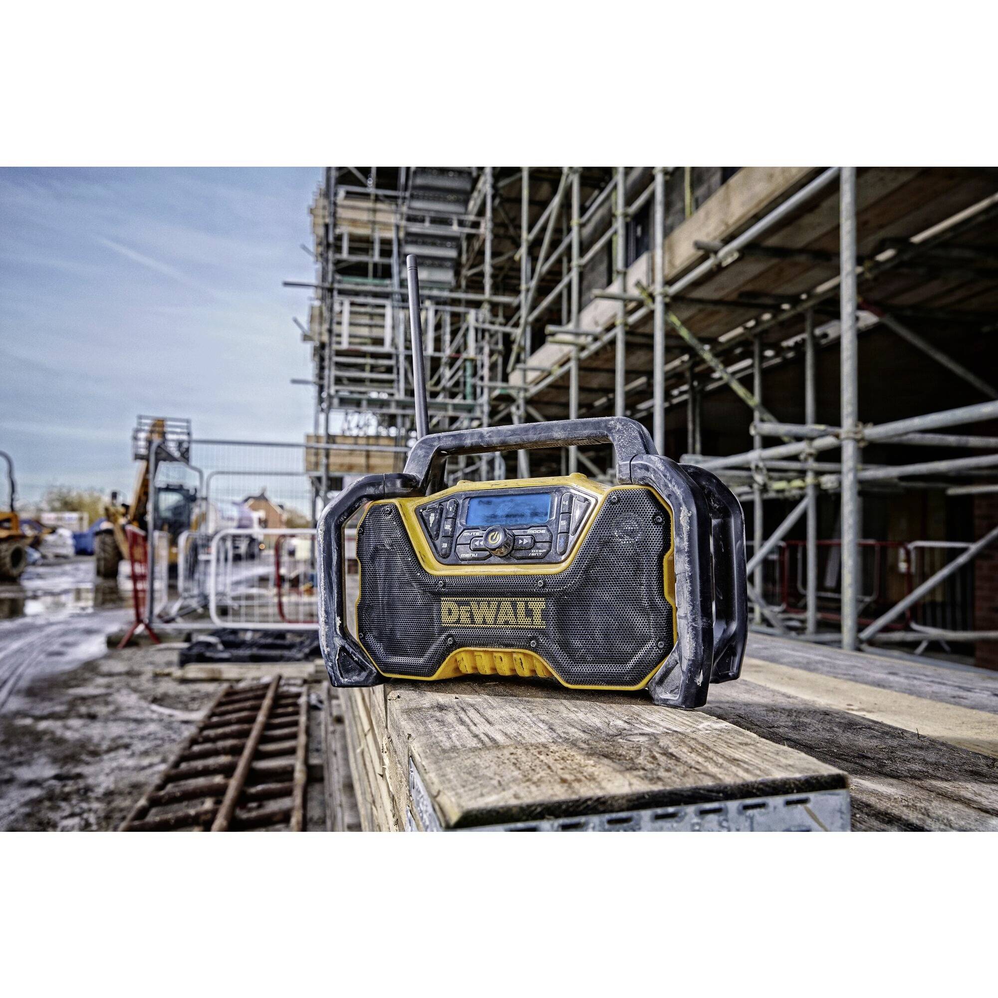 Byggarbetsplatsradio på ett träbord med en byggarbetsplats i bakgrunden, ställningar och byggmaterial, visar robust design under byggnadsförhållanden.