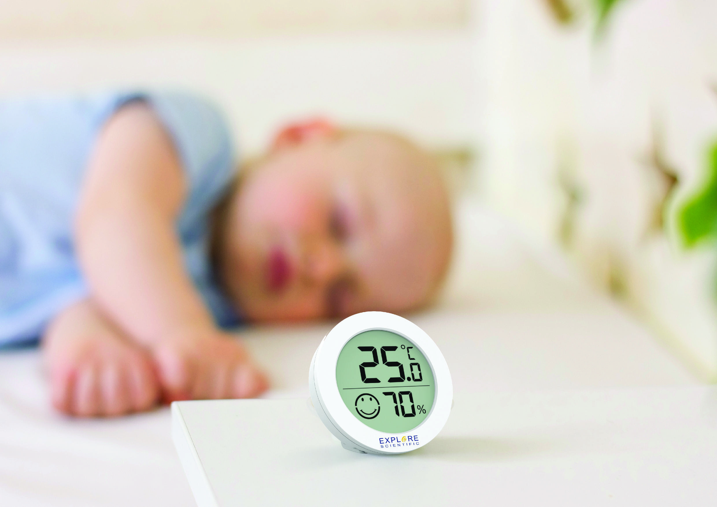 Ett sovande spädbarn i bakgrunden; i förgrunden ett digitalt termo-hygrometer som visar 25°C och 70% luftfuktighet.