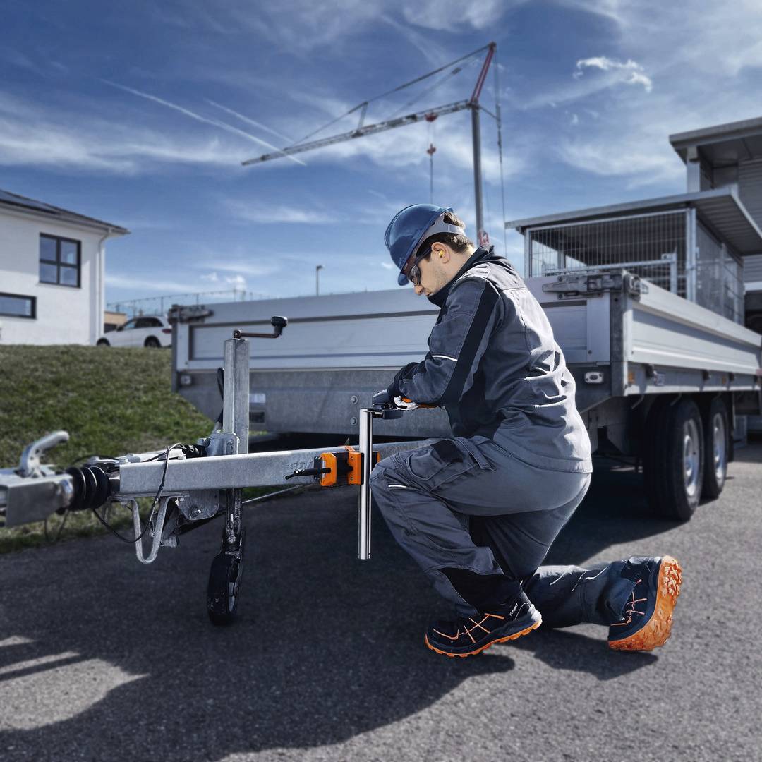 En byggnadsarbetare knäböjer bredvid en trailer och justerar en koppling. I bakgrunden syns byggnader och en kran.