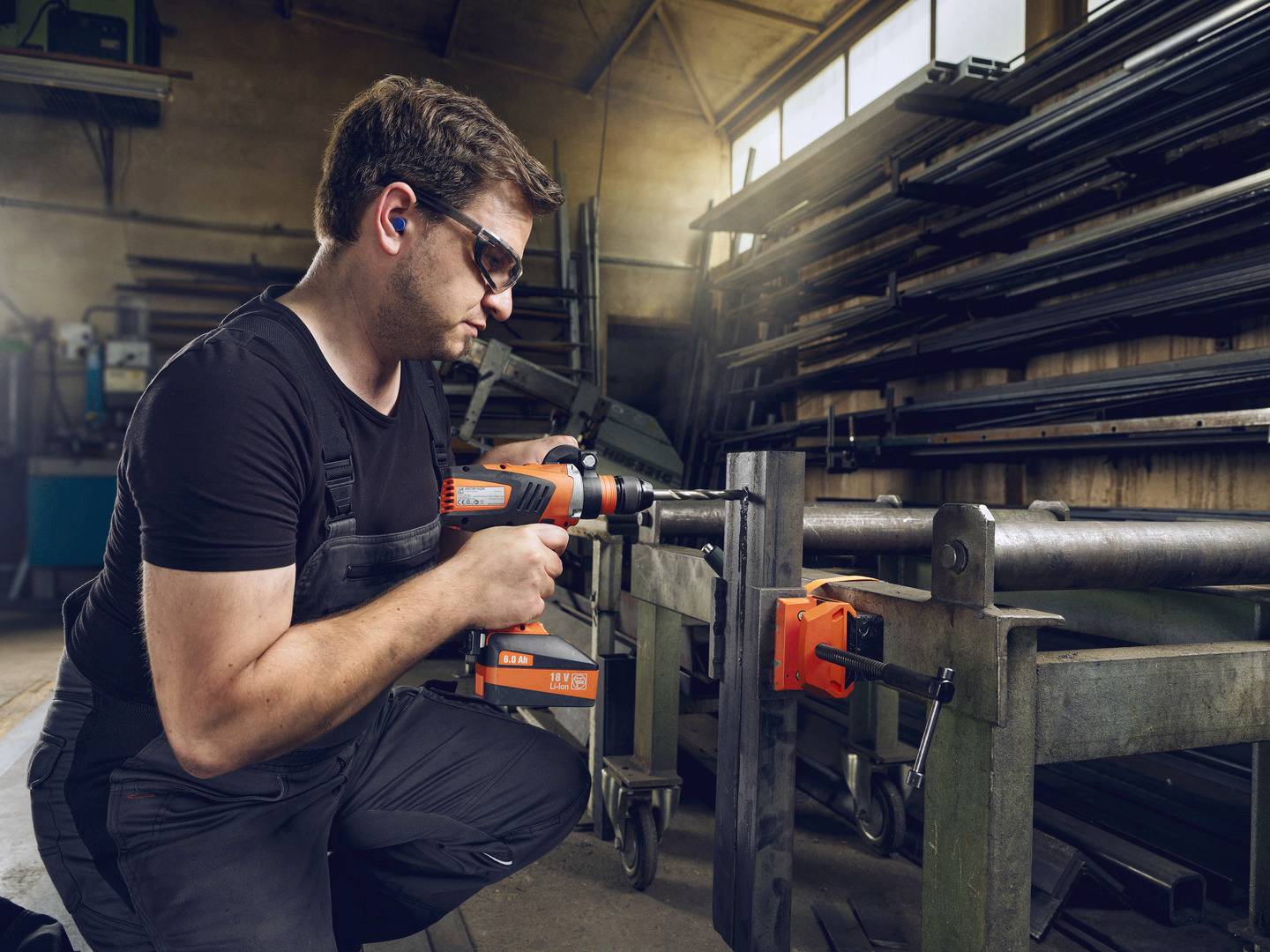 En man i arbetsklädsel borrar med en maskin i en verkstad. I bakgrunden syns metallhyllor och komponenter.
