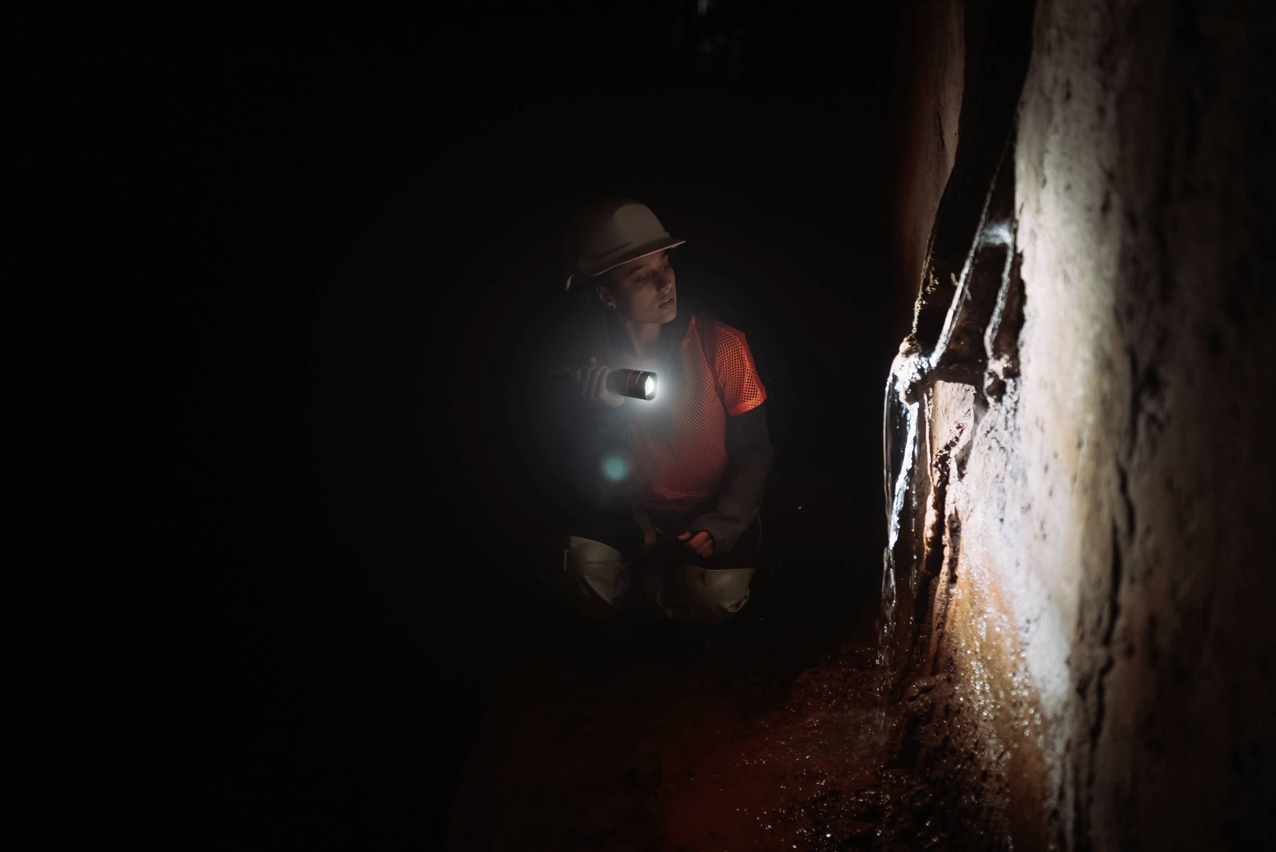En person i skyddsutrustning undersöker en mörk grotta eller tunnel med en ficklampa, fokuserad på en vägg.