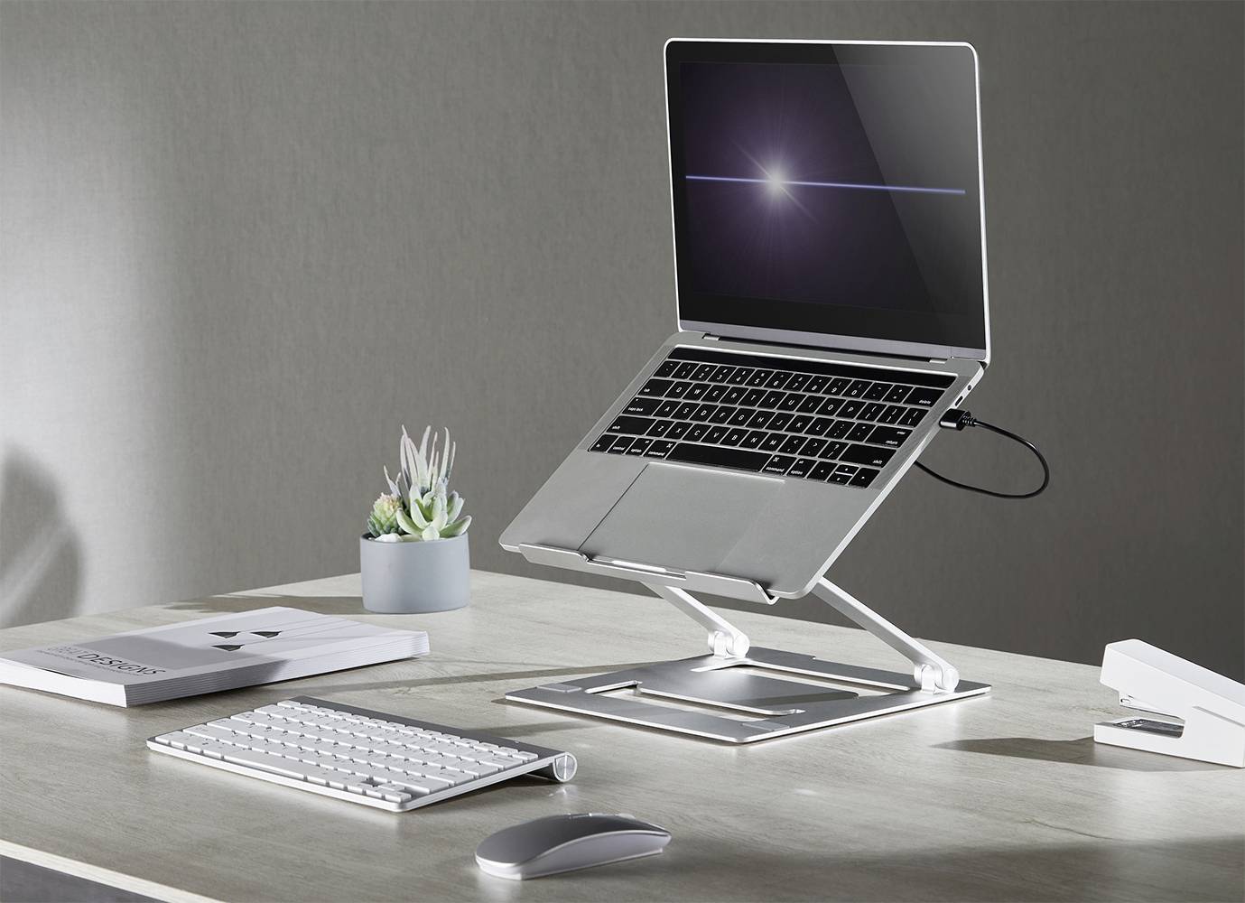 En bärbar dator på ett höjdjusterbart stativ på ett skrivbord med tangentbord, mus och krukväxt bredvid.