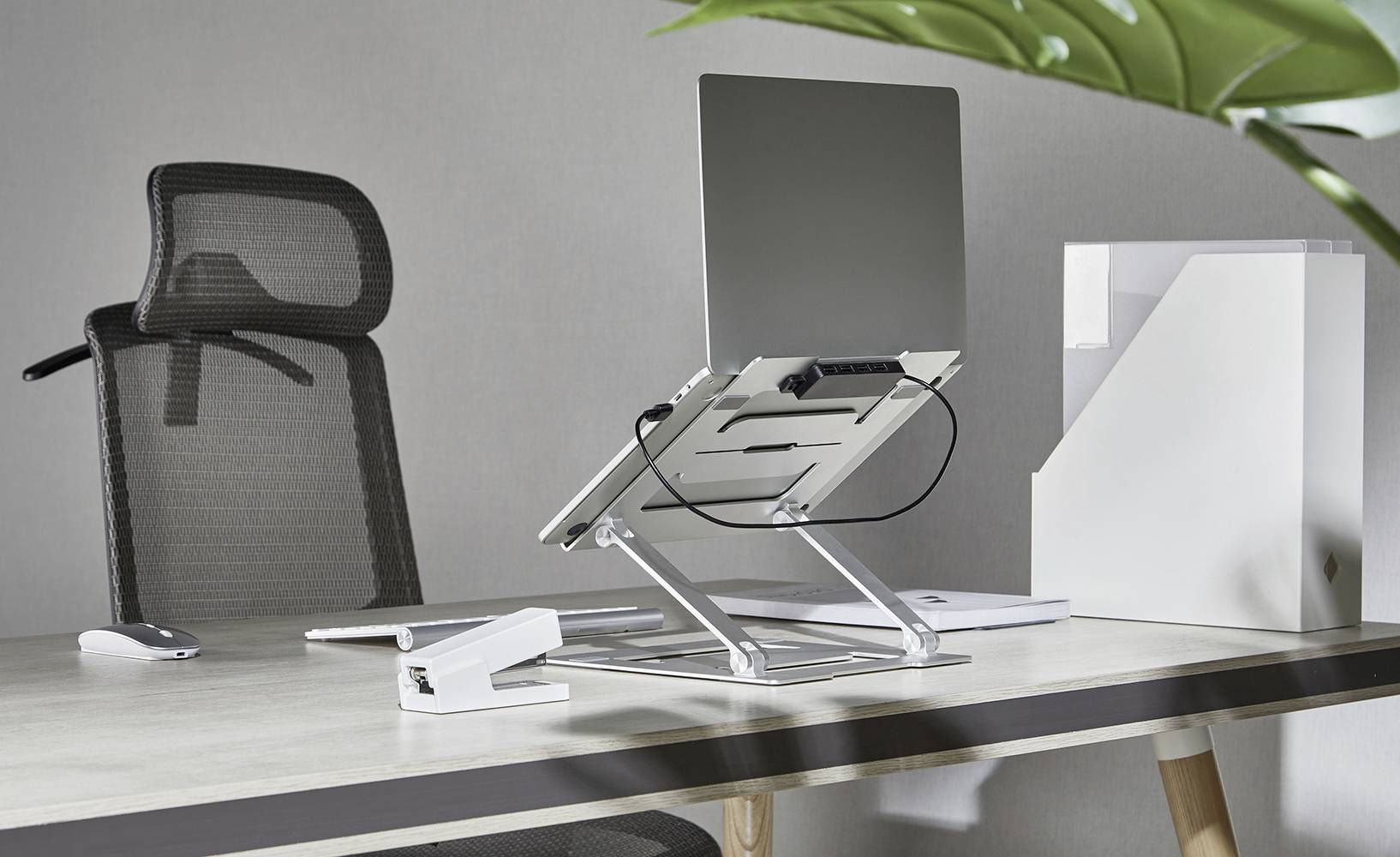 Modern skrivbord med ergonomisk stol, bärbar dator på stativ, dokumenthållare och häftapparat; perfekt för produktivt arbete.