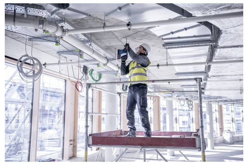 En byggnadsarbetare i en skyddsväst och hjälm använder en eldriven borrmaskin för att installera utrustning i taket på en modern byggnad under uppförande.