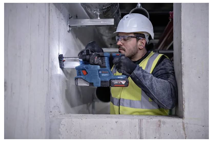 En byggnadsarbetare med hjälm och varselväst använder en slagborrmaskin på en betongvägg inuti en byggnad som är under uppförande.