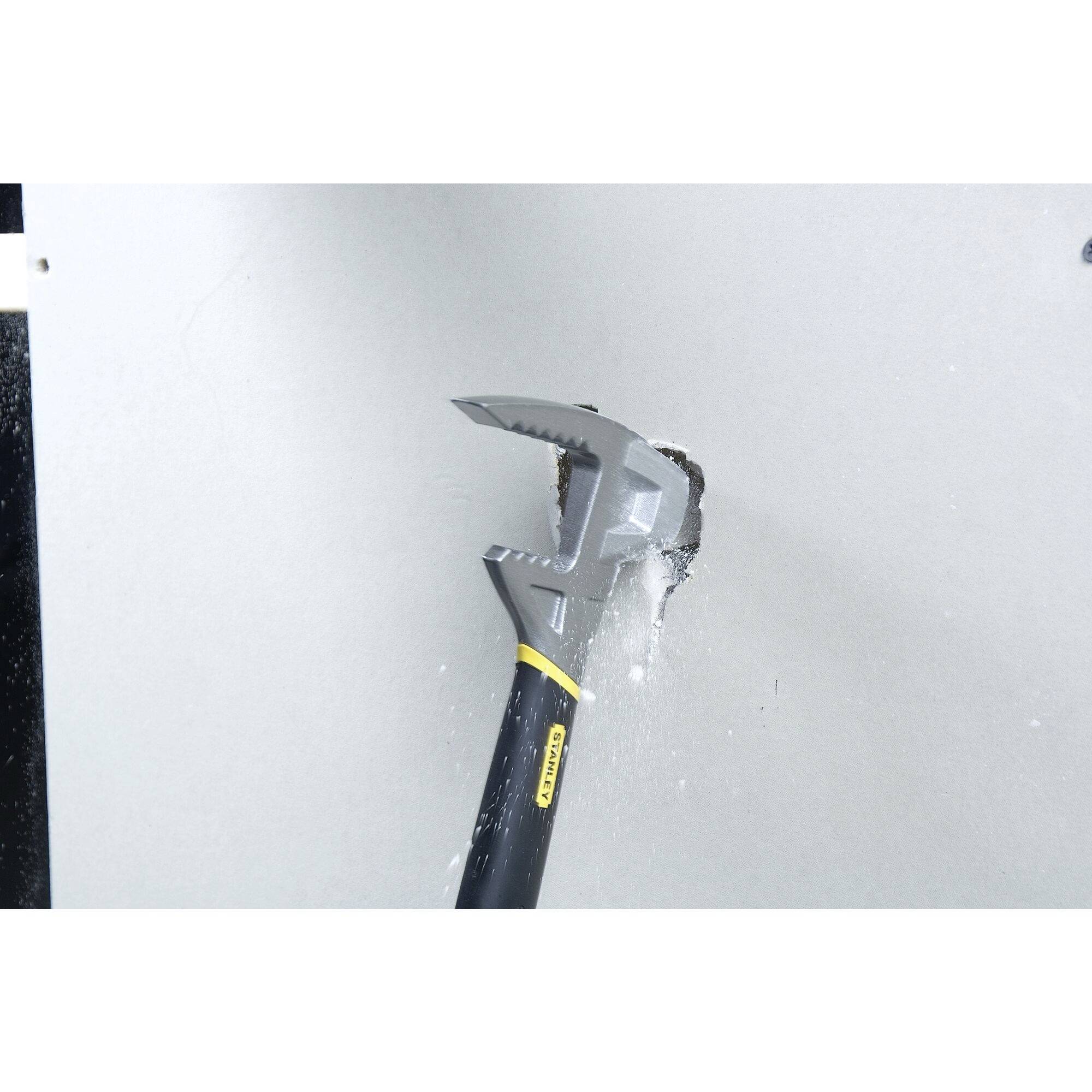 En hammare slår ett hål i en grå gipsskiva. Damm och skräp faller runt hålet. Hammaren är svart med gul accent.