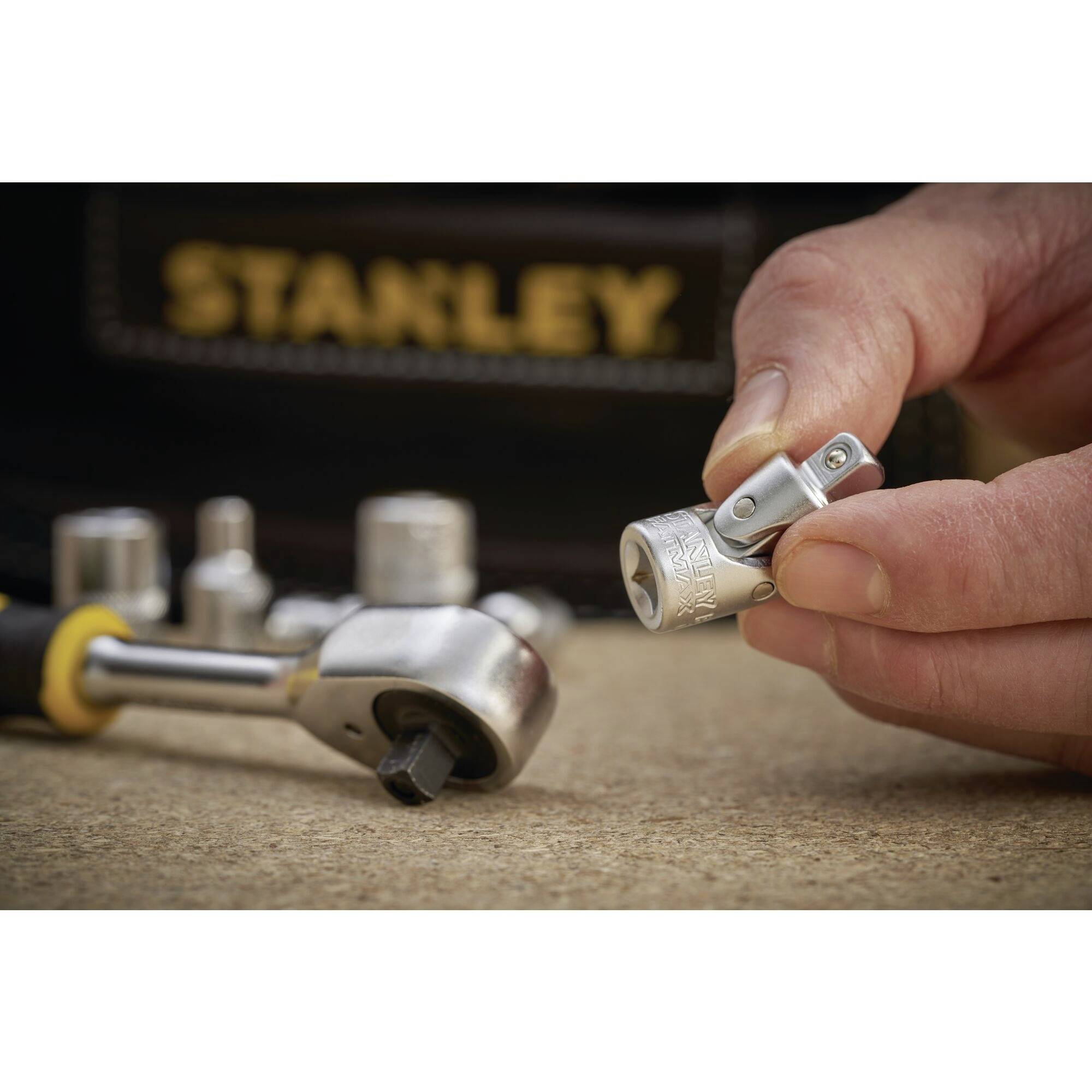 En hand håller en liten skruvmejseladapter. I bakgrunden syns fler verktyg och en väska märkt 'Stanley'.