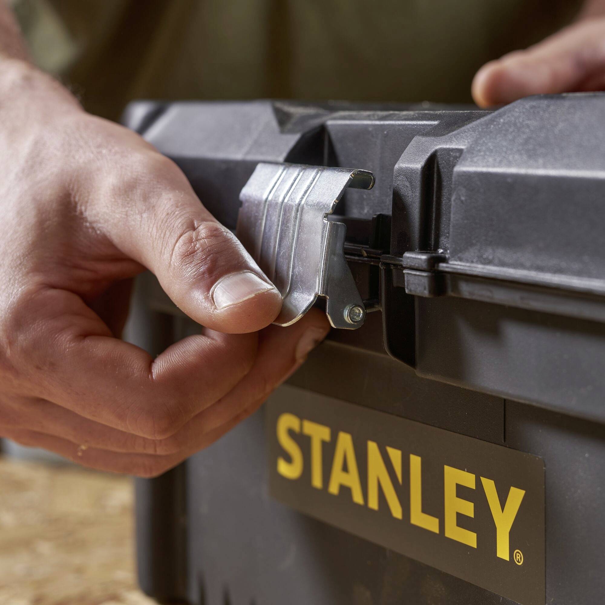 En hand öppnar låset på en verktygslåda från Stanley som står på en träyta.
