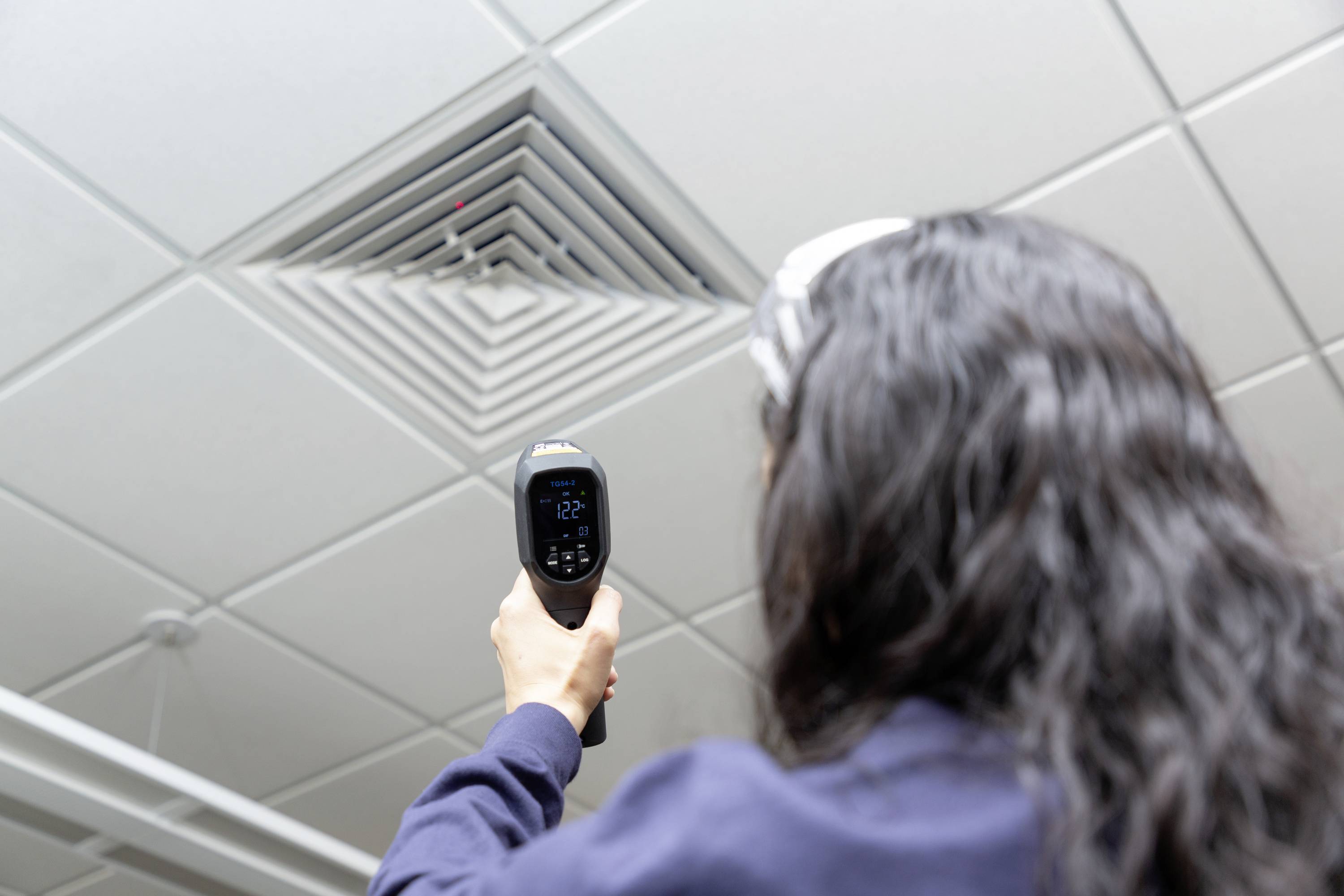 En person mäter temperaturen på ett ventilationsgaller i ett kontorstak med ett infraröd-termometer.