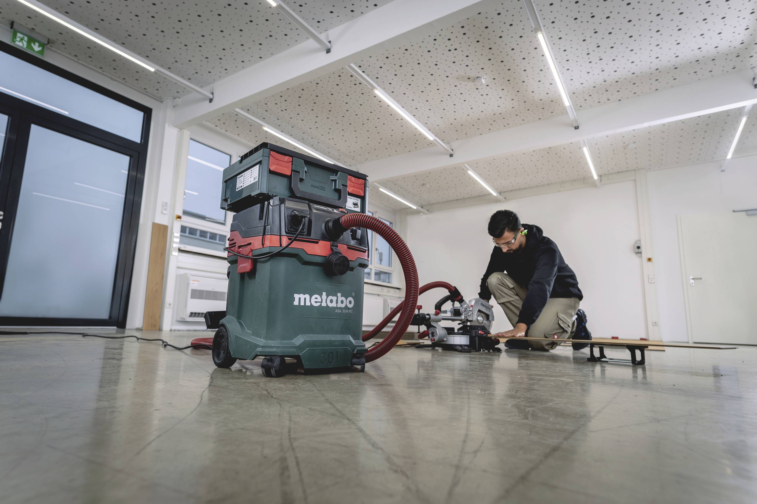 En person arbetar i ett rum med en Metabo industridammsugare i förgrunden. Rummet har ett nedpendlat tak med LED-belysning.