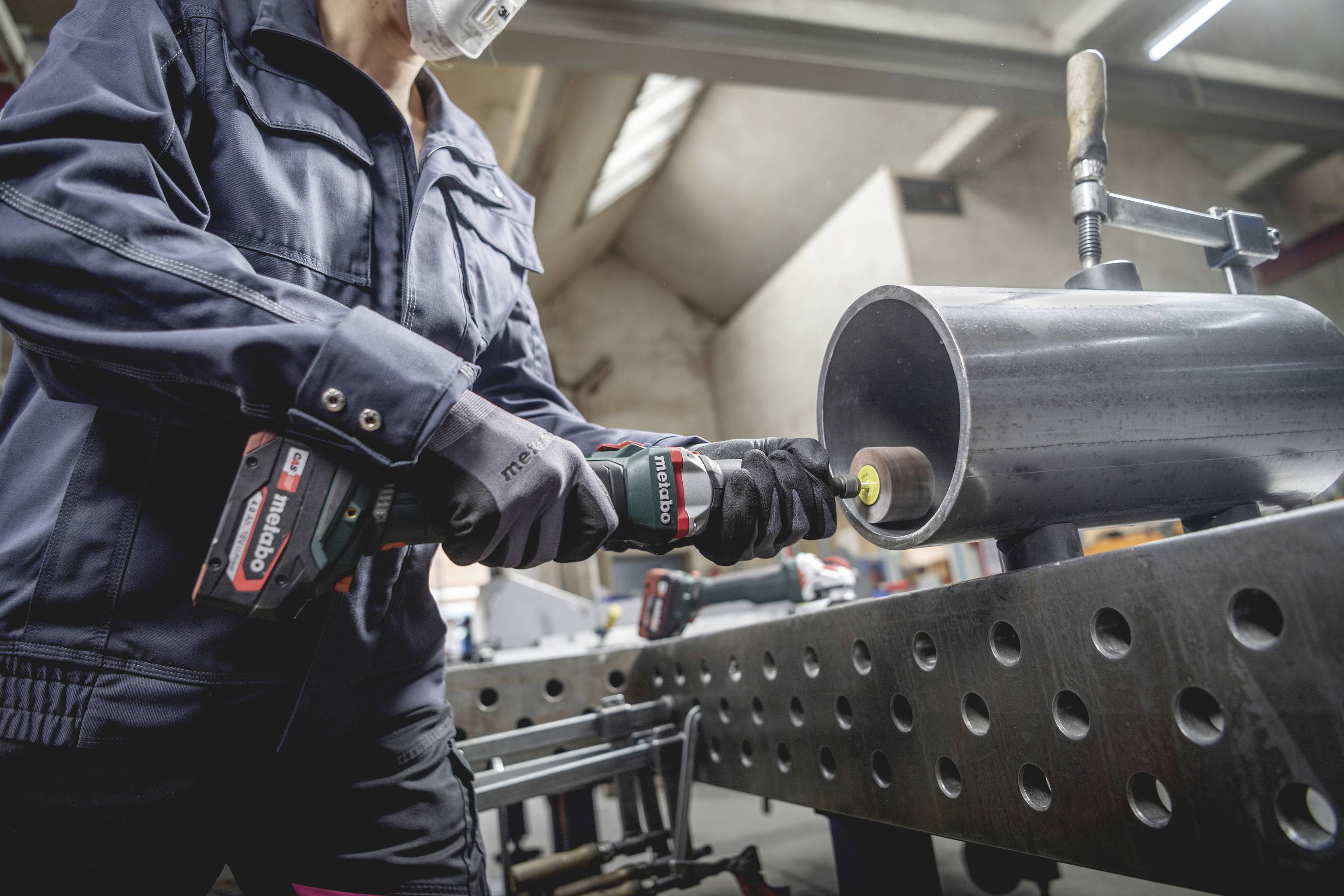 En person i arbetskläder slipar ett metallrör med ett handverktyg på ett verkstadsbord.