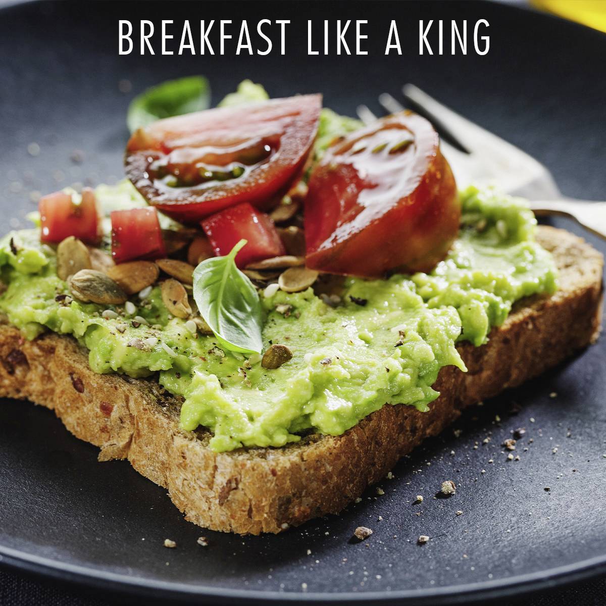 Ett rustikt bröd toppat med avokado, tomater och frön. I bakgrunden syns texten 'Frukostera som en kung'.