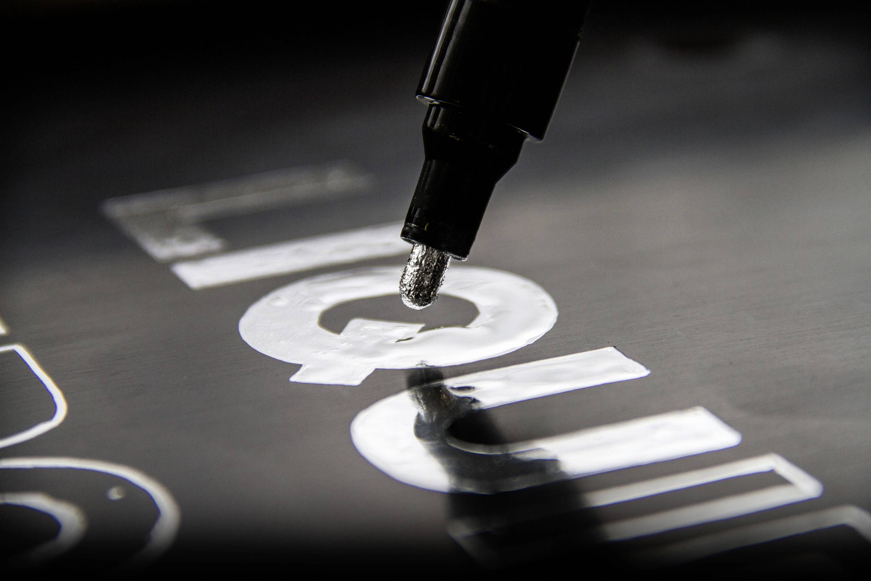 Närbild av en svart penna som målar vita bokstäver på en mörk yta. Fokus ligger på pennspetsen.