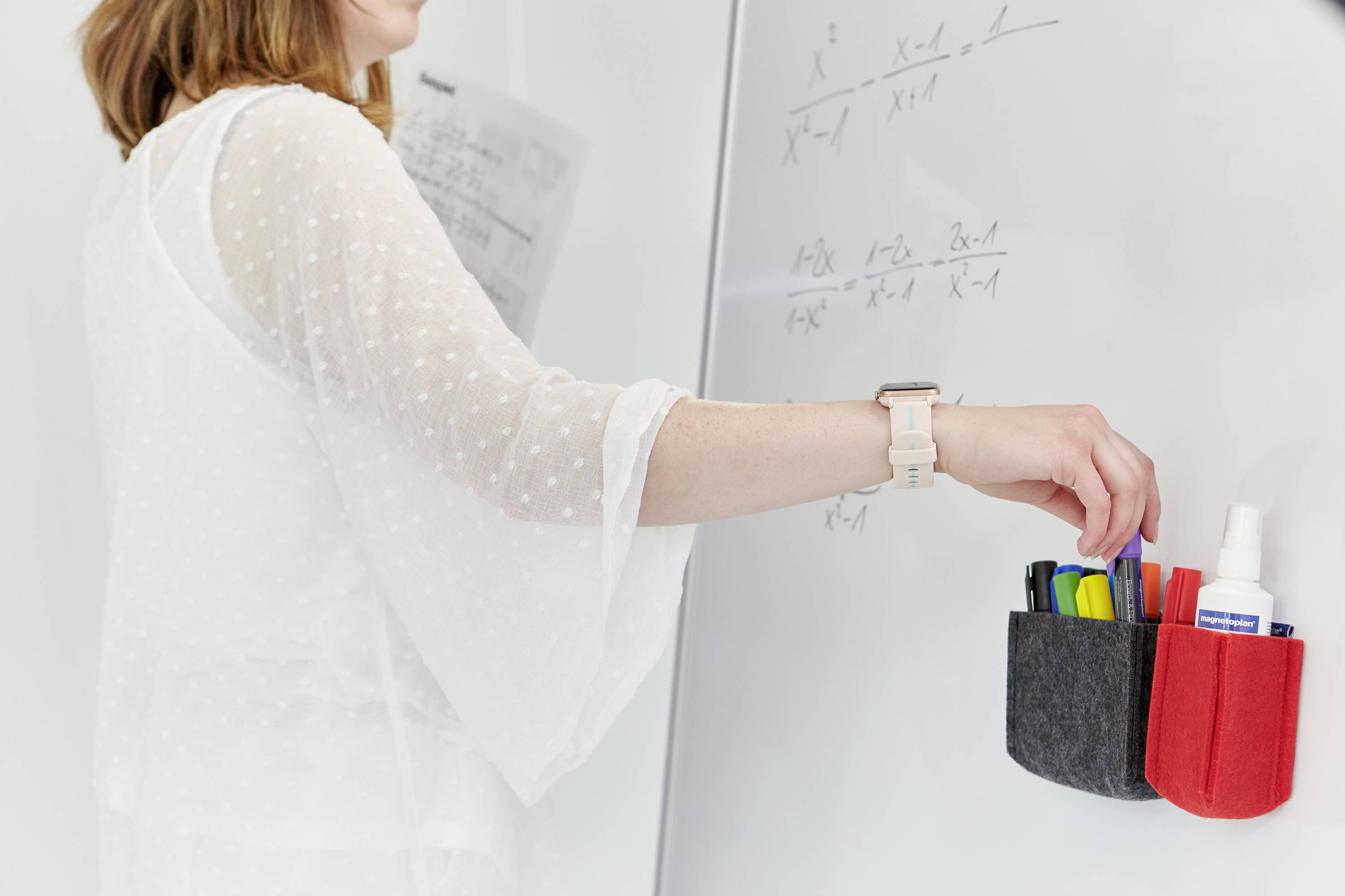 En person skriver matematiska formler på en whiteboard. I handen håller personen pennor och markörer från en väggställning.